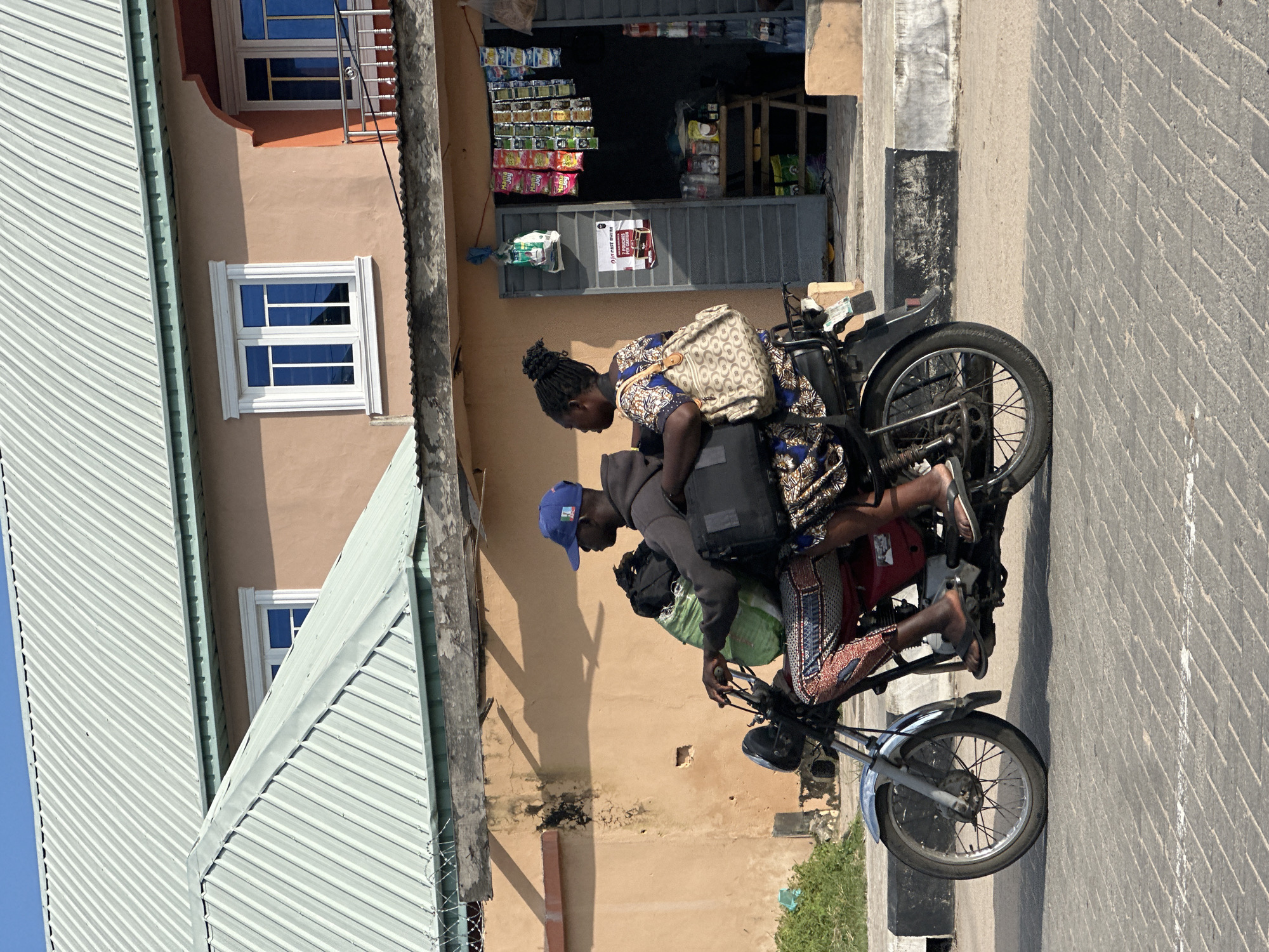 Two people are riding on a motorcycle. The person in the back, a woman, is wearing a patterned dress and a large patterned bag is slung over her shoulder. The person in the front, a man, is wearing a blue baseball cap and a gray hooded sweatshirt. He is holding onto the handlebars, and the woman is holding onto him. They appear to be traveling together.
The motorcycle is carrying several bags. A green bag is strapped to the back of the motorcycle, and a dark bag is behind the man. The motorcycle is on a paved road. The scene appears to be in a commercial area. In the background, there is a storefront with shelves stocked with various goods. A sign on the storefront reads "FAST PHARM".
The sun is shining, suggesting it is daytime. The shadows cast by the people and the motorcycle indicate a sunny day, likely mid-morning or afternoon. The overall impression is a bustling street scene in a town or city in Nigeria. The people seem to be focused on their journey, and there are no strong emotional cues visible. The buildings in the background have a distinctly Nigerian architectural style, with corrugated metal roofs and plastered walls.
