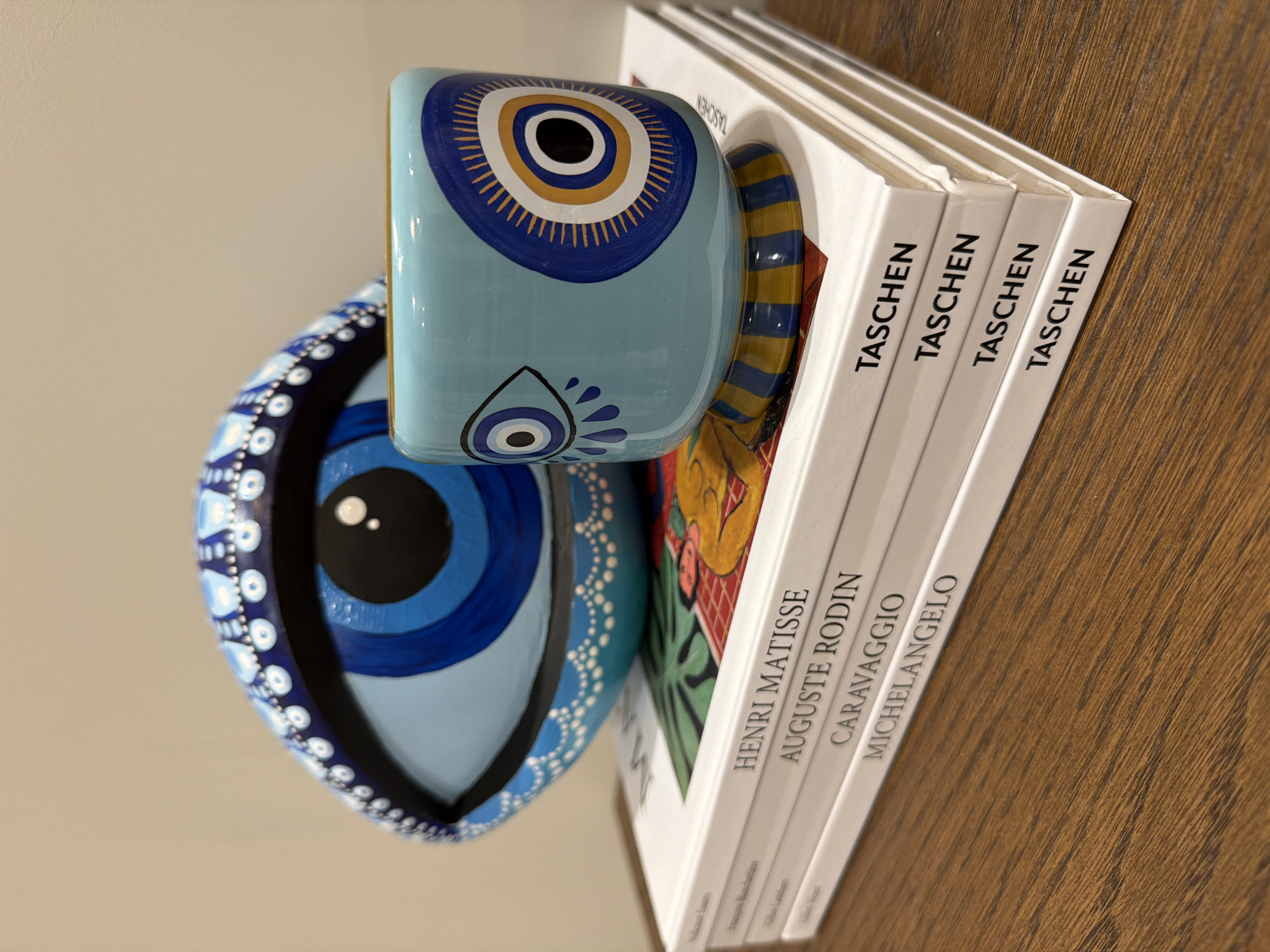 The image shows a close-up of several items: a stack of Taschen art books, a light blue ceramic mug, and a blue and white painted ceramic bowl. The books, stacked on a wooden surface, appear to be about Henri Matisse, Auguste Rodin, Caravaggio, and Michelangelo. The mug, placed on top of the books, is decorated with an eye design. The bowl, also featuring an eye design, rests next to the mug. No people are visible. The wooden surface has a visible grain. There is no indication of a specific location in Yerevan, Armenia.