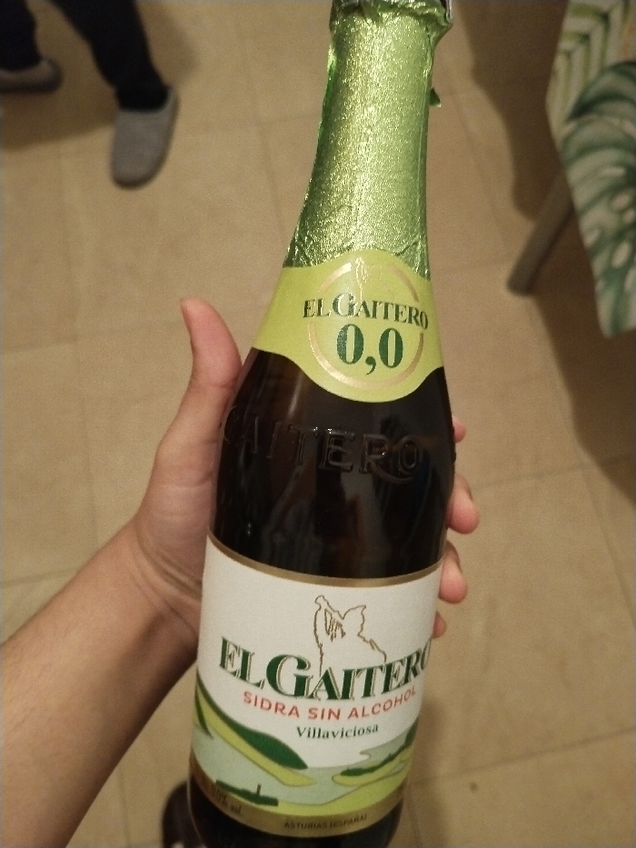 A hand holds a bottle of El Gaitero Sidra Sin Alcohol, a non-alcoholic cider. The bottle has a green foil top and a label with the "El Gaitero" logo and "0,0" indicating no alcohol content. The label depicts a figure playing a bagpipe. "Sidra Sin Alcohol" and "Villaviciosa" are also printed. The lower part of the label features a stylized landscape. Tile flooring and a glimpse of furniture are visible in the background.