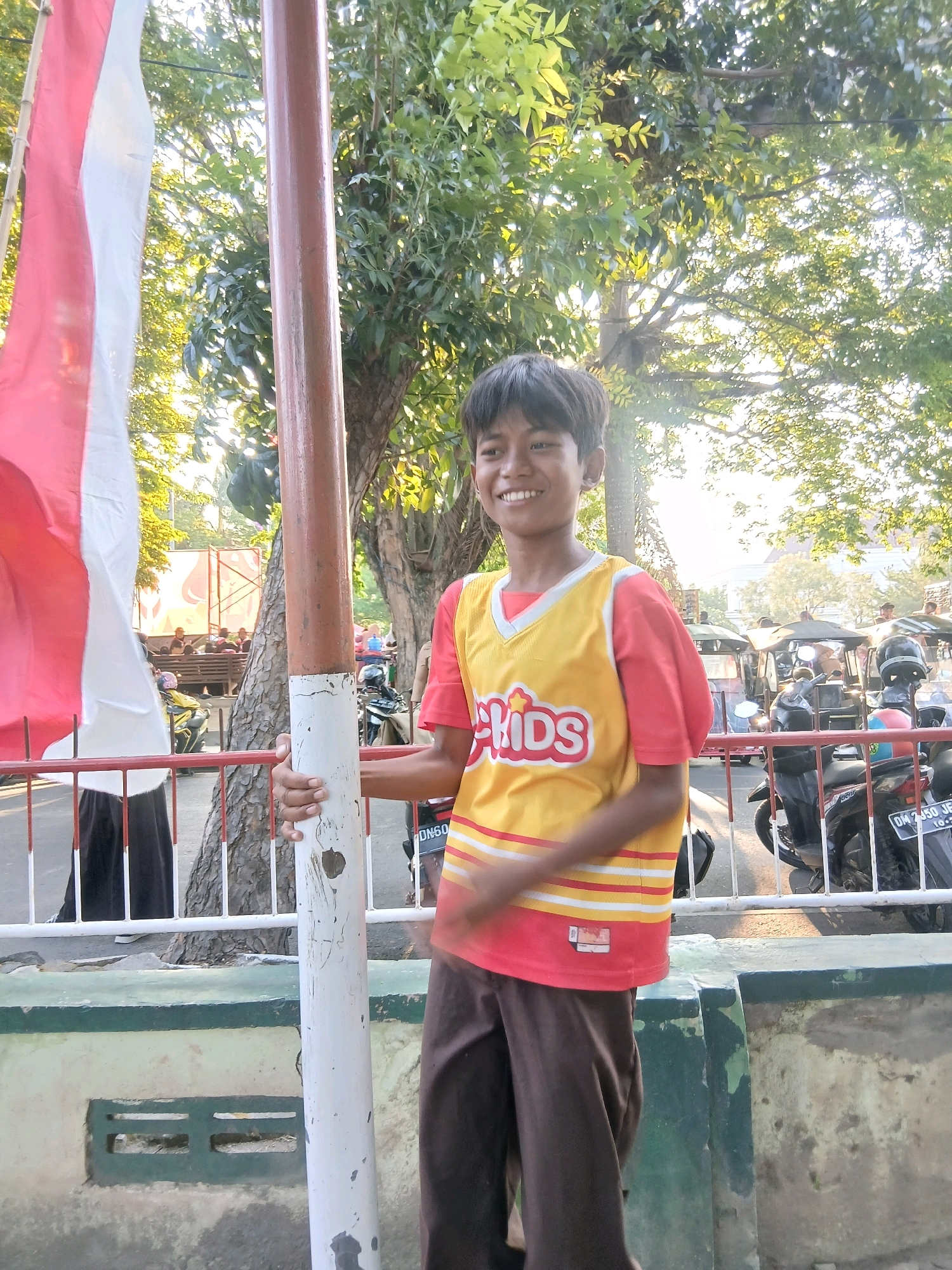 A young Indonesian boy with dark hair, wearing a red t-shirt with a yellow basketball-style jersey over it and brown pants, stands outside holding a white pole that has a reddish-brown section near the top. The jersey has "KIDS" printed on it in red and white letters with a yellow outline. To the boy's left, a red and white Indonesian flag hangs from a pole. In the background, trees with green foliage are visible, with sunlight filtering through the leaves. Behind the boy, a white metal fence separates him from a street area where several motorbikes and what appear to be three-wheeled vehicles are parked. The ground in front of the boy consists of a concrete barrier painted green on top and a rough concrete surface below. A partially visible building with a white facade and red accents can be seen in the distance. The scene appears to be in an outdoor public space, possibly a park or a roadside area, in the city of Gorontalo, Indonesia.