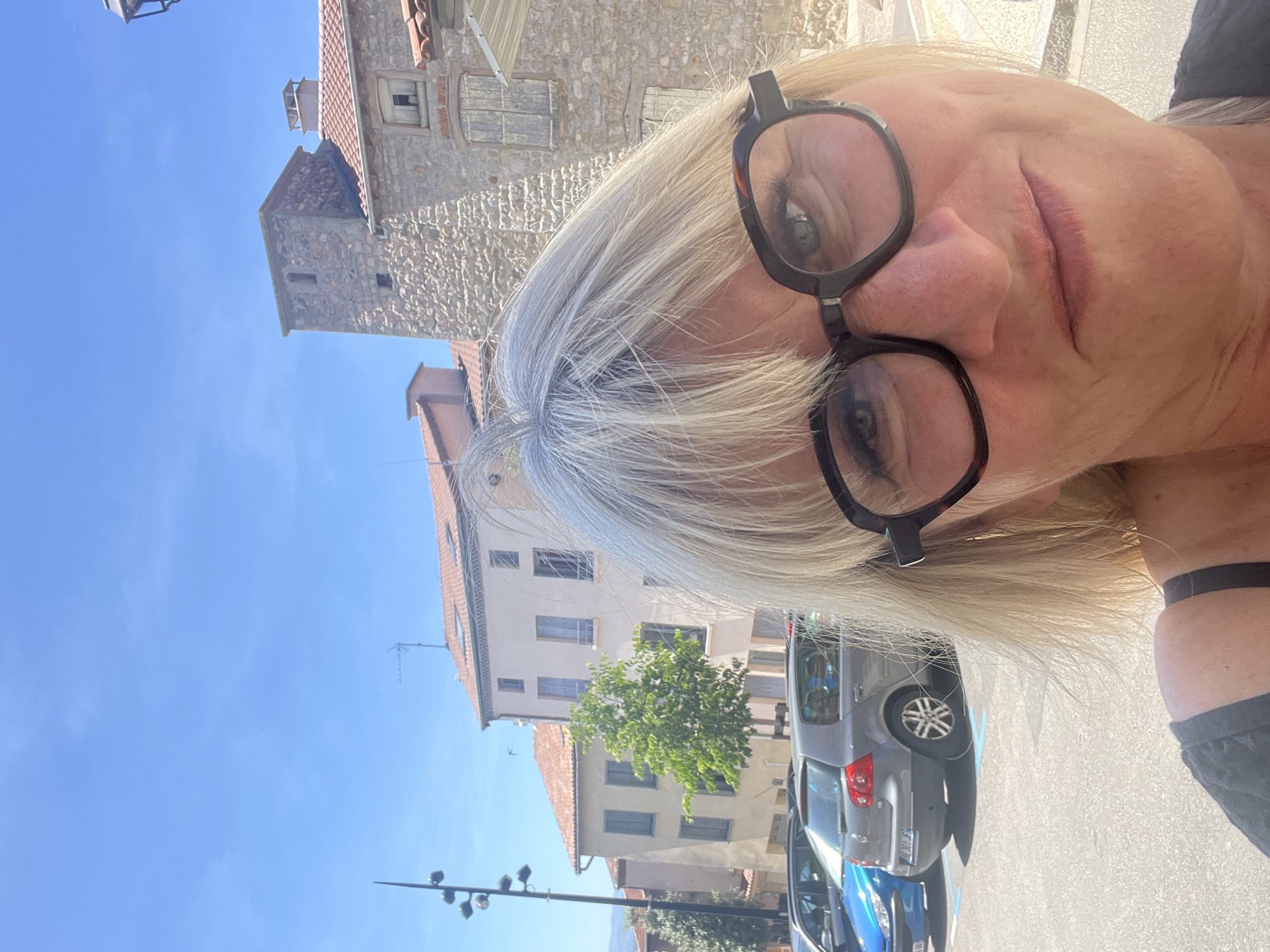 In Saint-Galmier, France, on a sunny day with a bright blue sky, a woman with blonde hair and dark-rimmed glasses takes a selfie. She is partially visible, with her face and upper chest in the foreground. Behind her, a street scene unfolds. Several cars are parked on the right side of the image, including a blue car and a grey Volkswagen Golf. The buildings in the background are made of stone and plaster, with terracotta roofs. A tall, stone tower with a small window stands out against the sky. A street lamp with multiple lights is visible on the left side of the frame. The overall impression is that of a quaint European town on a clear day.