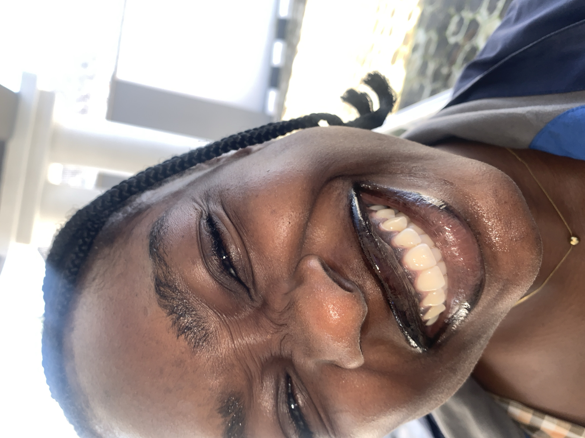 The image is a close-up, angled shot of a young Black woman's face, taken from a low perspective. She is smiling widely, with her eyes squinted shut and her nose scrunched. Her lips are full and glossy with dark lipstick, revealing bright white teeth. Her skin is smooth and appears dewy. She has braided hair, with one braid visible over her shoulder. She is wearing a plaid shirt underneath a gray and blue jacket, and a thin gold chain with a small heart pendant is visible around her neck. The background is blurred, but suggests an outdoor setting with bright light, possibly sunlight filtering through structures, hinting at a contemporary urban or semi-urban environment. There is no discernible indication of the specific location within Gura Top, Nigeria.