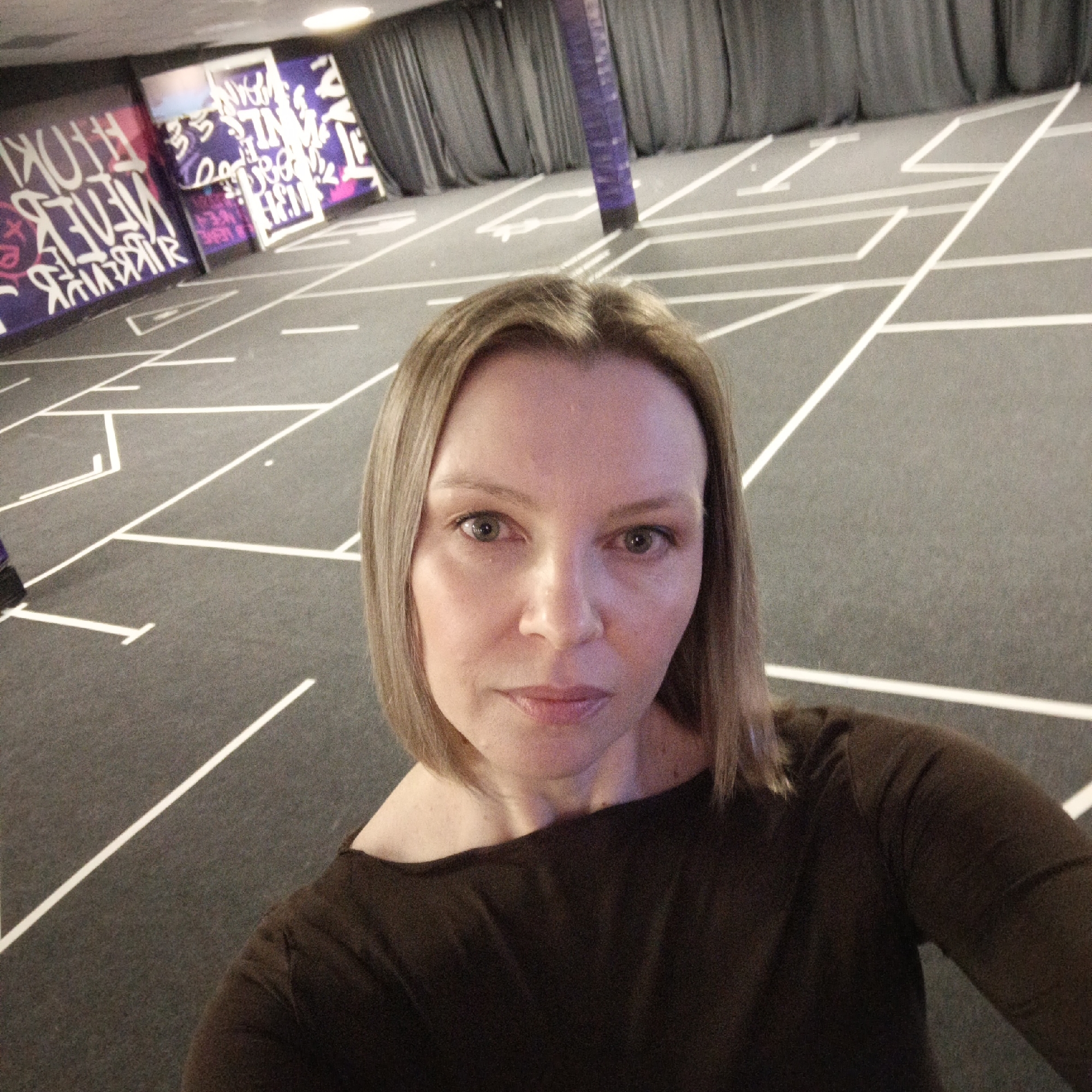 A woman with shoulder-length blonde hair and a dark brown, off-the-shoulder top is taking a selfie. The setting appears to be an indoor space with a dark gray floor marked with a white geometric pattern resembling a hopscotch layout or a labyrinth. In the background, there are wall panels with graffiti-style artwork in vibrant colors, and gray curtains or drapes. A purple pillar is also visible in the background. The location is possibly a game room or interactive installation in the city of Chaikovsky, Russia, based on potential association of the graffiti panels with local art scenes or specific landmarks there.