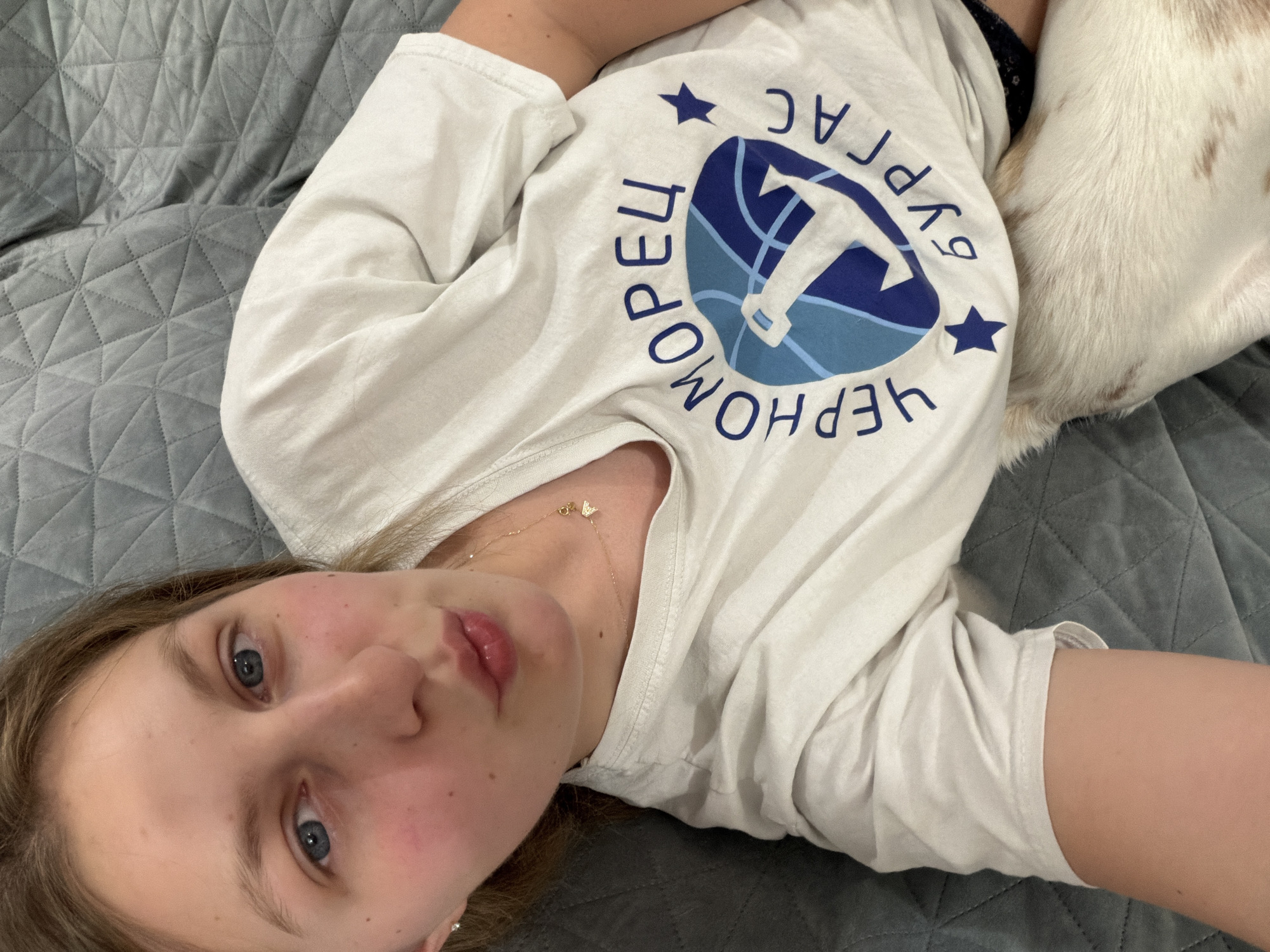 A young woman with blonde hair and blue eyes poses for a selfie from a reclined position on a grey quilted surface. She is wearing a white t-shirt with a blue circular logo depicting a lighthouse and the text "ЧЕРНОМОРЕЦ БУРГАС" in Cyrillic. Her lips are slightly pursed, and she is wearing a delicate gold necklace. Part of a white and brown dog is visible near her lower torso. The background is a grey quilted fabric, likely a sofa or bed. The scene appears to be indoors, possibly in a residence in Burgas, Bulgaria, given the text on the t-shirt.