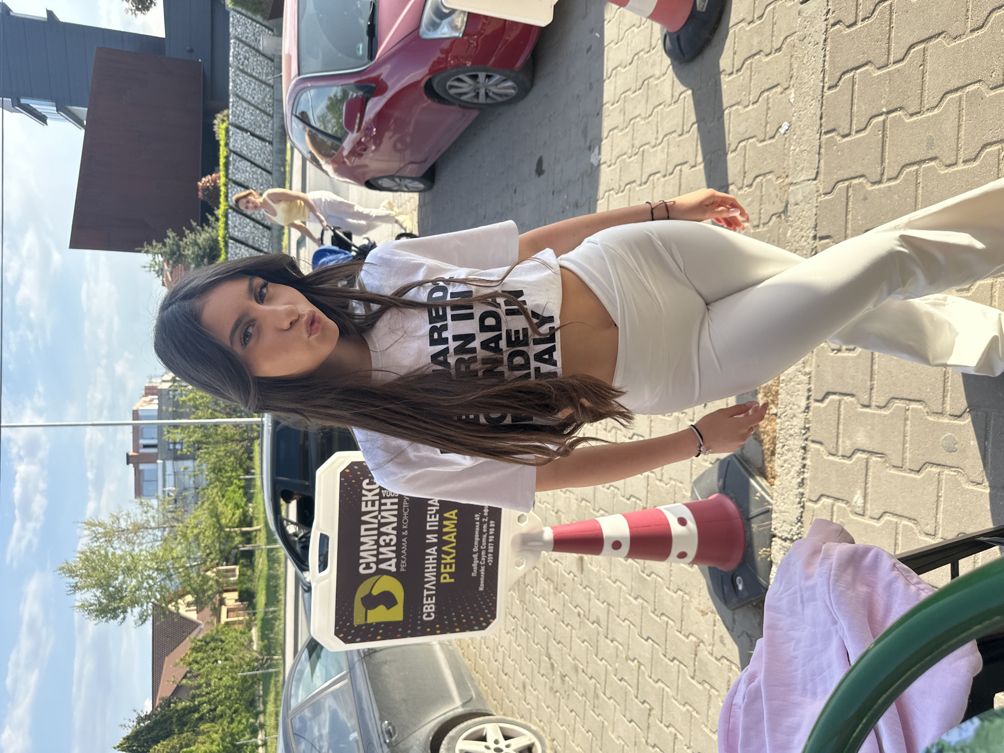 An outdoor, sunlit, full-length shot captures a young woman posing on a paved surface in Plovdiv, Bulgaria. She has long, dark brown hair and is looking directly at the camera with a slight pout. She is wearing a white cropped t-shirt with black text, partially visible as "ARED RN IN NADA DE IN TALY," likely referring to clothing branding. She pairs this with matching high-waisted white pants. On her right wrist, she wears several thin dark bracelets.
The setting appears to be a commercial or residential area. In the background, a red car is parked, and a darker car is partially visible to the left. Further back on the left, another person is seen walking with a blue baby stroller. The ground is paved with interlocking concrete pavers, and clear shadows suggest bright sunshine, indicating daytime. A red and white striped traffic cone is situated near her right side.
A prominent sign stand to the left of the woman displays Bulgarian text. It reads "СИМПЛЕКС ДИЗАЙН РЕКЛАМА & КОНСТРУ" (SIMPLEX DESIGN ADVERTISING & CONSTRUCTION) and "СВЕТЛИННА И ПЕЧА РЕКЛАМА" (LIGHT AND PRINT ADVERTISING). Below this, an address is provided: "Пловдив, Остромила 49, Комплекс Саут Сити, ет. 2, офи" (Plovdiv, Ostromila 49, South City Complex, floor 2, office), along with a phone number "+359 887 98 98 89." In the immediate foreground on the right, a pink garment is draped over a green chair or railing. Buildings and trees line the background, contributing to an urban outdoor scene.