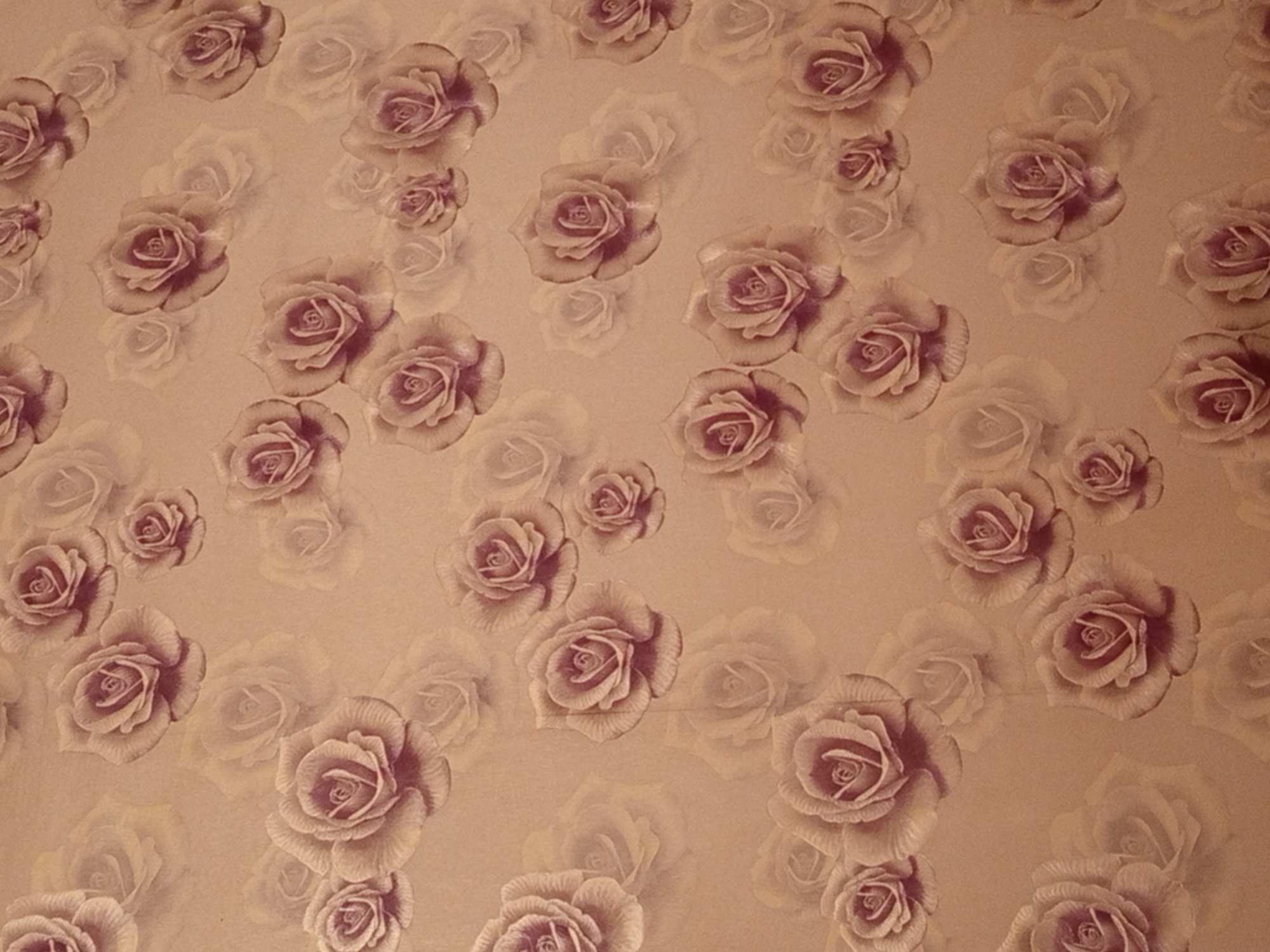 The image displays a seamless, repeating pattern of stylized roses in various sizes and shades of pink and brown against a textured, muted background that appears to be wallpaper or fabric. The roses are depicted with detailed petals, and some are shown in full bloom while others are in bud form. The overall aesthetic is vintage and romantic, with a subtle, soft lighting that gives the impression of a gentle glow. There are no people, animals, or specific objects that would indicate a location in Ylay-Talaa, Kyrgyzstan, or any other geographical place. The image is solely a decorative pattern.