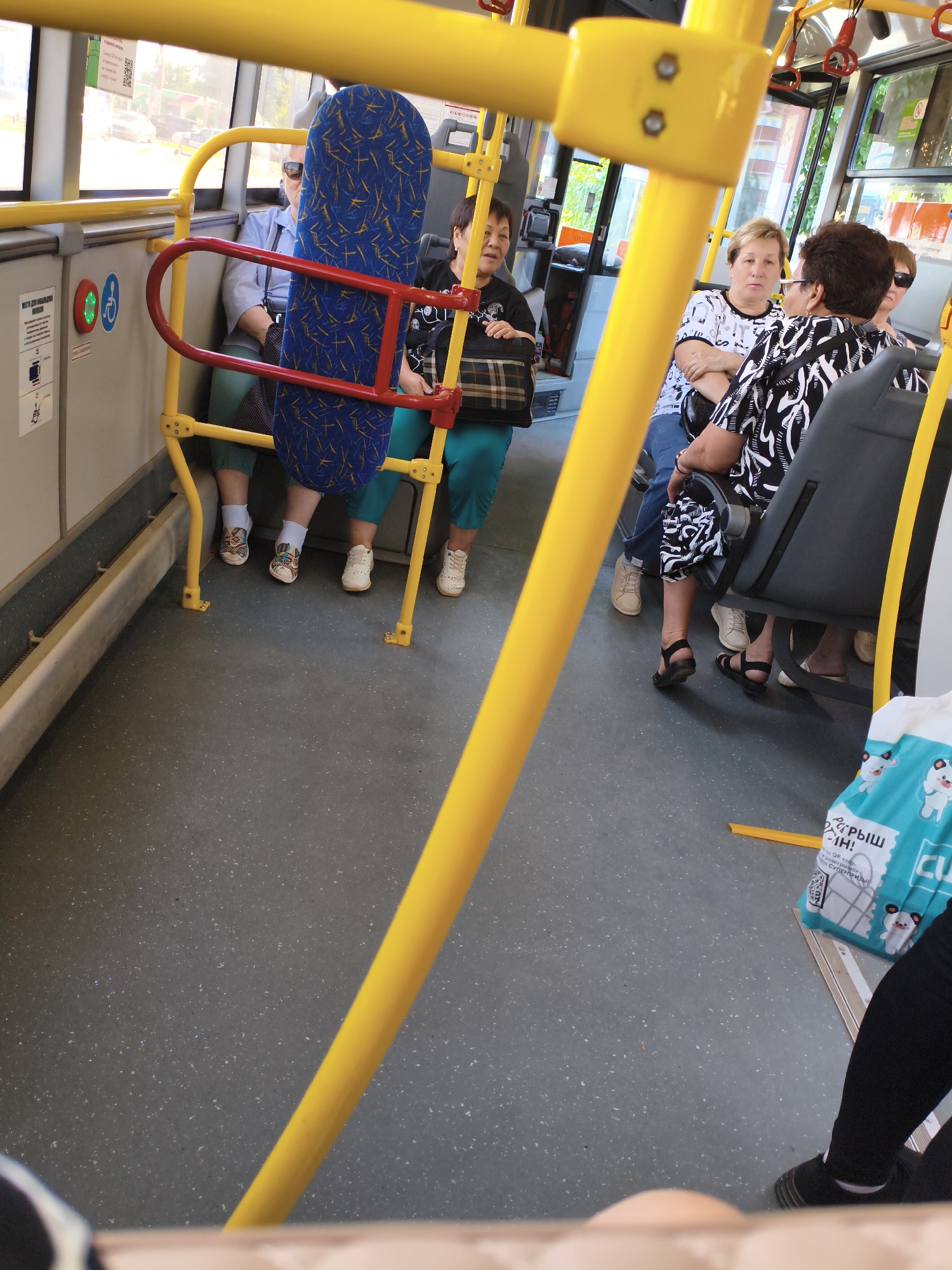 The image shows the interior of a public bus, likely in Orenburg, Russia, based on the provided information. The scene is viewed from a low angle, looking upwards and towards the front of the bus. Yellow metal poles and bars are prominent throughout the frame, indicating the bus's internal structure and safety features.
In the foreground, partially obscured by a yellow pole, is a blue seat with a patterned fabric. To the left of this seat, a woman in a blue shirt and green pants is seated, facing away from the camera. Next to her, another woman, also in green pants, is seated and facing towards the camera. She is wearing a black and white patterned top. Further down the aisle, another woman in a black and white patterned top and dark pants is seated.
The floor of the bus is a speckled gray material. Various objects are visible, including a blue and white patterned bag with images of cartoon animals and text in Russian, placed on the floor near the aisle. Red handrails are attached to the yellow poles. On the wall of the bus, there are signs, including one with pictograms of a person and a wheelchair, suggesting it is an accessible vehicle. The windows of the bus allow natural light to enter, and blurred outdoor scenery is visible through them, suggesting movement.