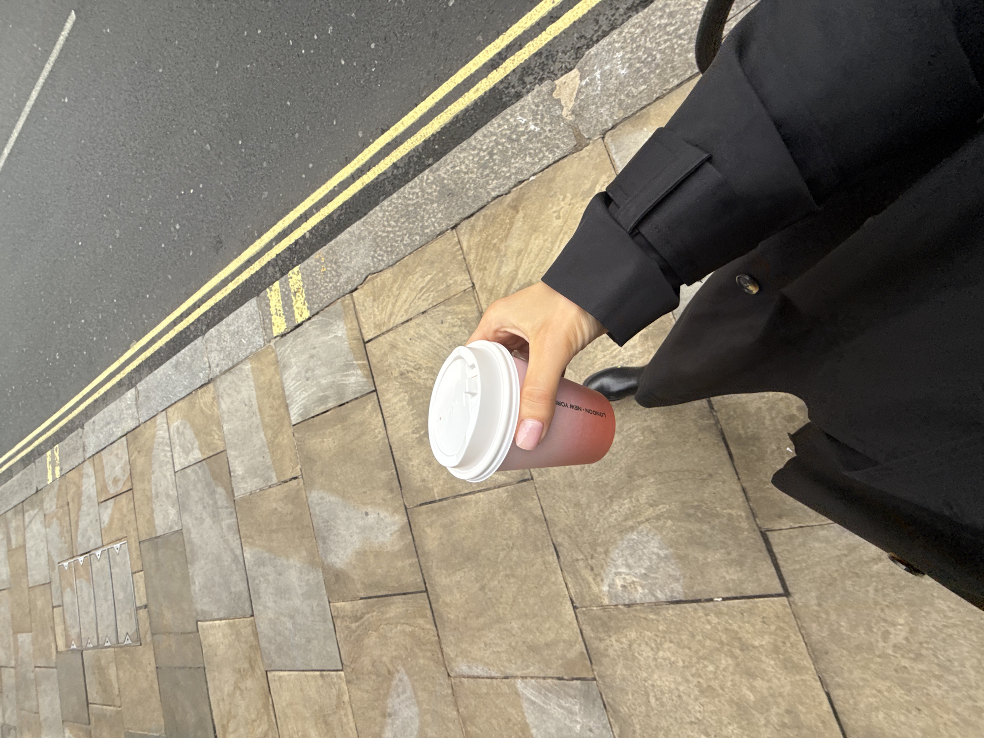 The image provides an overhead view of a person's right arm and hand holding a clear, reddish-pink beverage cup with a white lid. The person is wearing a black long-sleeved garment, possibly a coat or jacket. The setting is an urban street, featuring an asphalt road to the left with prominent double yellow lines, indicating parking restrictions common in the UK. Adjacent to the road is a curbstone, followed by a pavement composed of rectangular, light-colored stone slabs, some of which show darker or lighter patches, possibly from dampness.
Visible text on the cup, oriented upside down from the viewer's perspective, clearly reads "LONDON NEW YORK". This, coupled with the double yellow lines, strongly suggests the location is in London, United Kingdom. The time of day appears to be daytime, with soft, diffused lighting and no strong shadows, possibly indicating an overcast sky. The activity depicted is simply the person holding a drink while positioned next to a road and pavement.