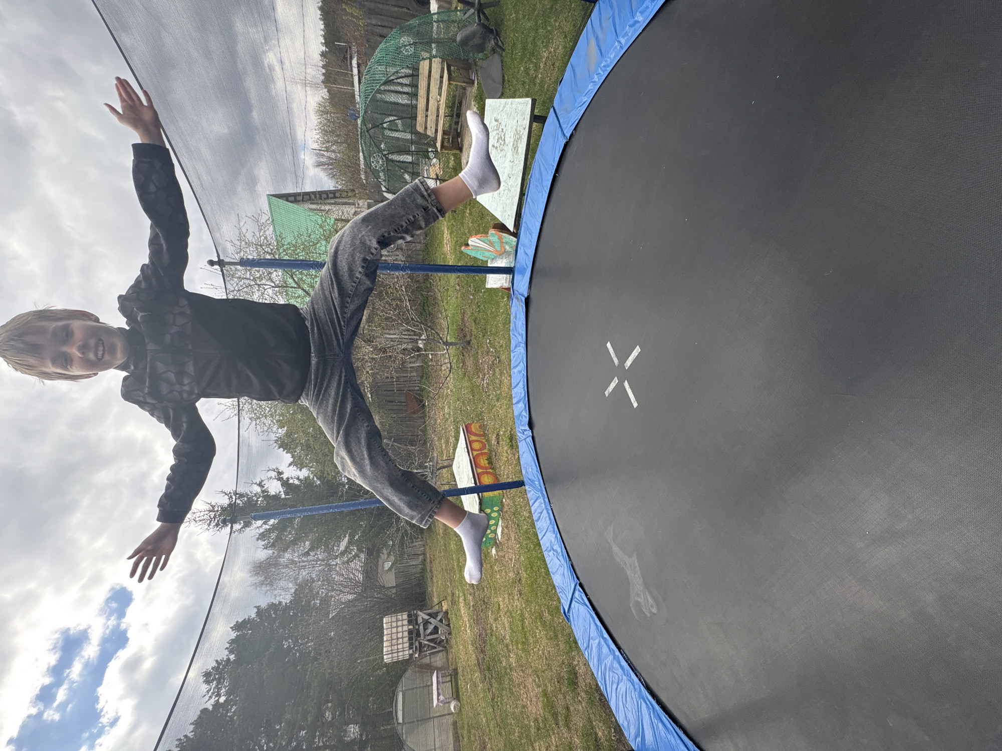 A child is in mid-air, seemingly falling off a trampoline. The child is wearing a black jacket, grey jeans, and white socks. The trampoline is round with a blue safety pad and a black jumping surface. The background shows a yard with grass, various plants, a greenhouse, and a structure resembling a bench or small building. The sky is cloudy. The location is likely in a rural or suburban area, potentially near Chaikovsky, Russia.