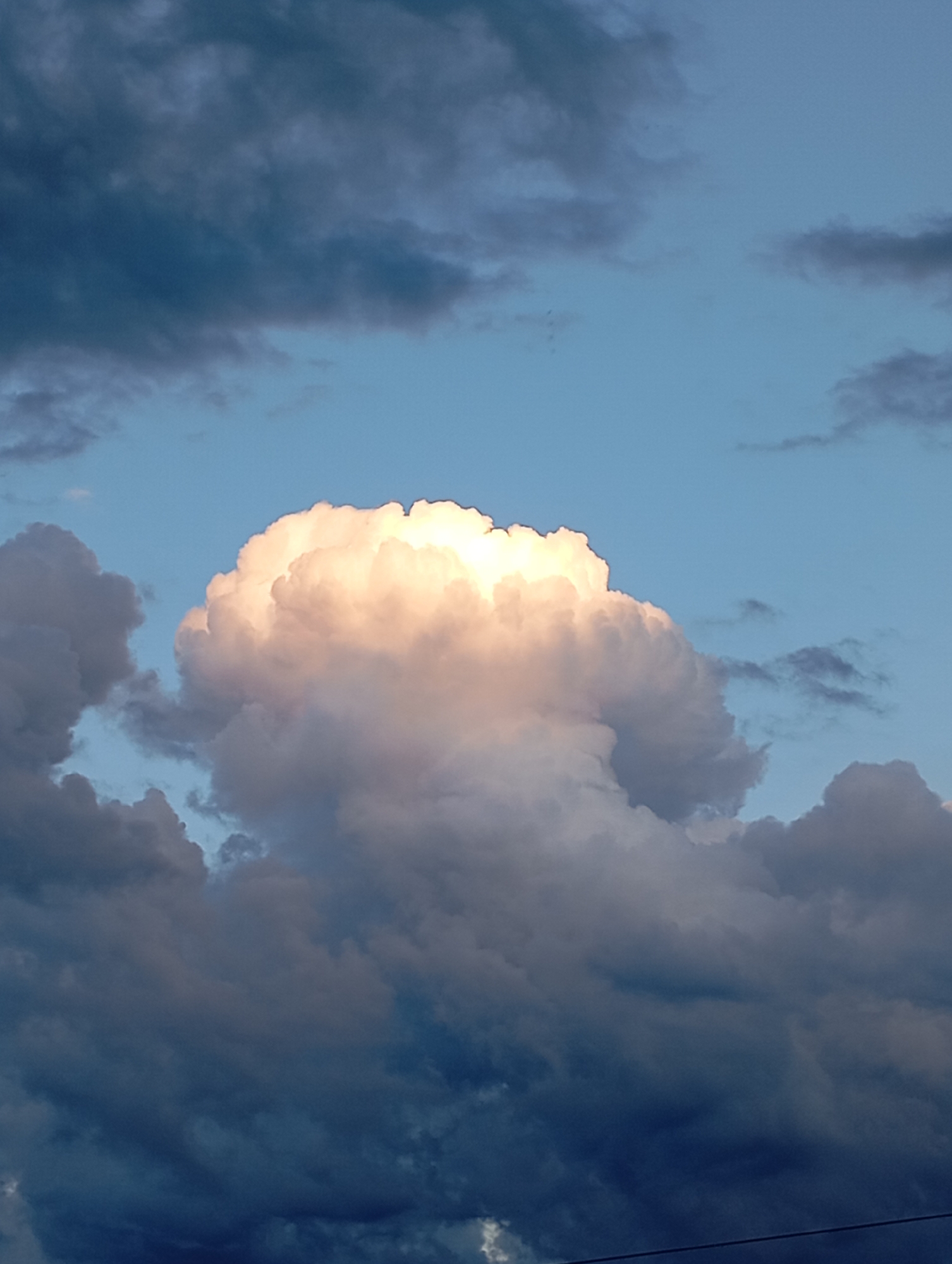 The image depicts a cloudy sky with a large, bright cumulus cloud illuminated by sunlight in the center. Darker, heavier clouds surround the central formation. The lower portion of the image shows a dark, dense cloud bank, suggesting impending weather changes. The scene is a sky view and does not contain any identifiable elements related to the city of Ryabki, Russia.