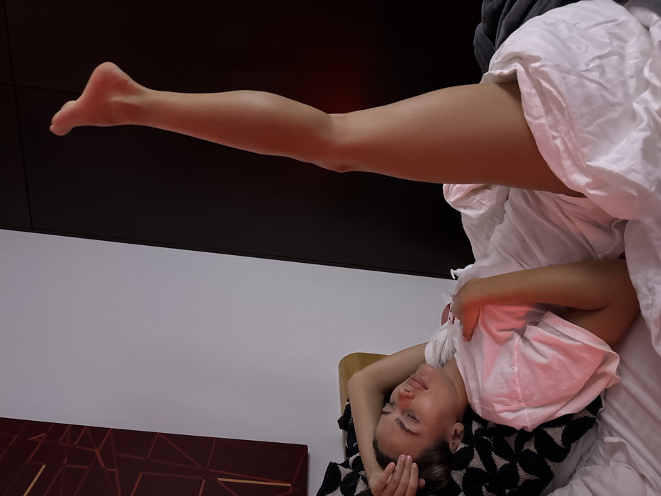 A woman lies on her back in bed, covered by a white sheet and a black-and-white patterned blanket. Her head rests on a light-colored headboard, and her eyes are closed. One leg is extended straight up in the air, foot pointed. The background consists of a white wall on the left and a dark, possibly brown, panel above. A reddish glow is cast upon the skin and bedding. A painting with geometric shapes is partially visible in the lower-left corner.
