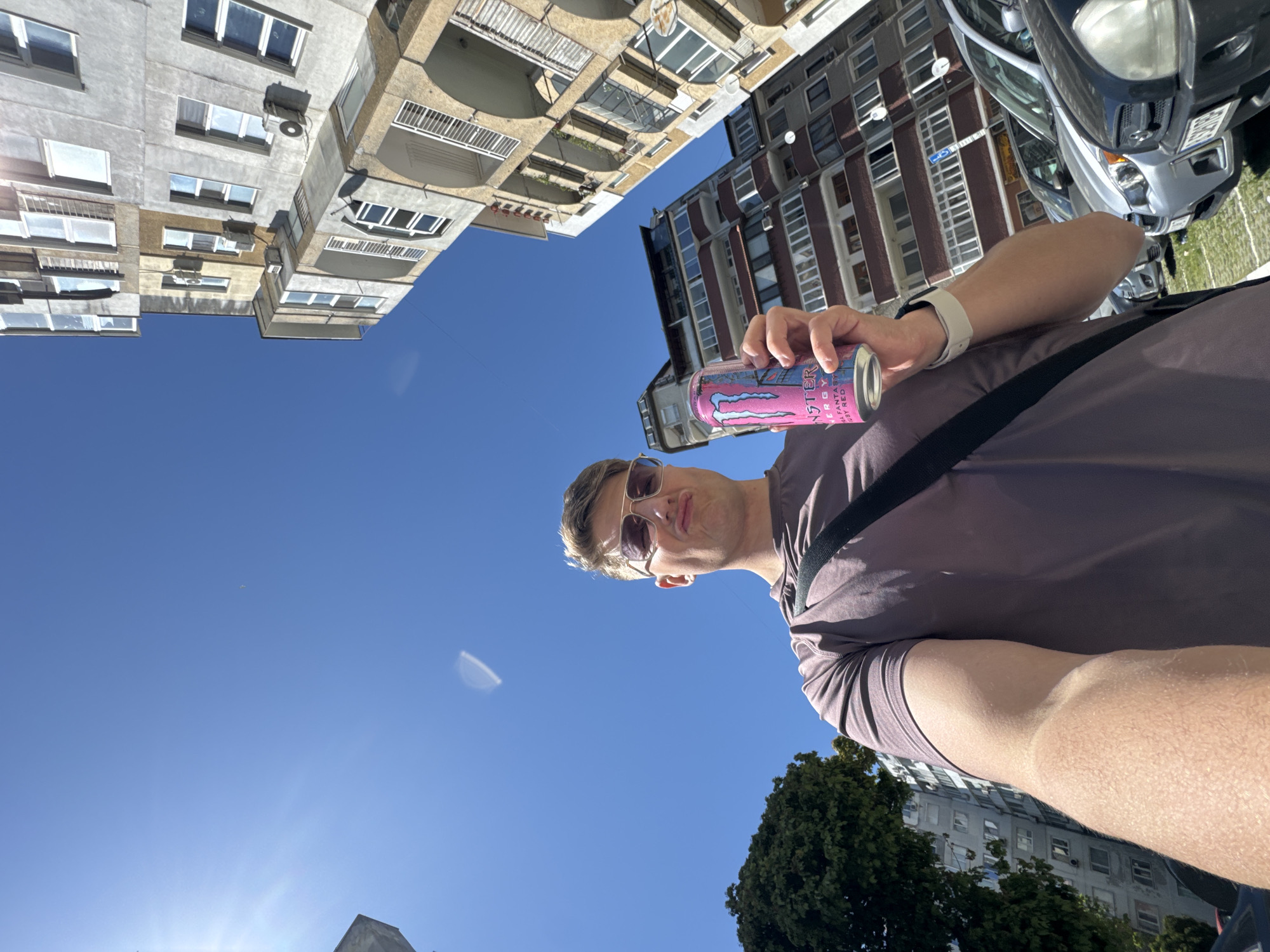 A man is holding a can of pink Monster energy drink, against a bright blue sky. He is wearing sunglasses and a short-sleeved shirt, with a strap crossing his chest. Buildings are visible in the background, a mix of older, Soviet-era apartment blocks and more modern constructions. Trees are also visible in the background, partially obscuring some of the buildings. Based on the architecture and the presence of Soviet-era buildings, it is likely that the location is Varna, Bulgaria.