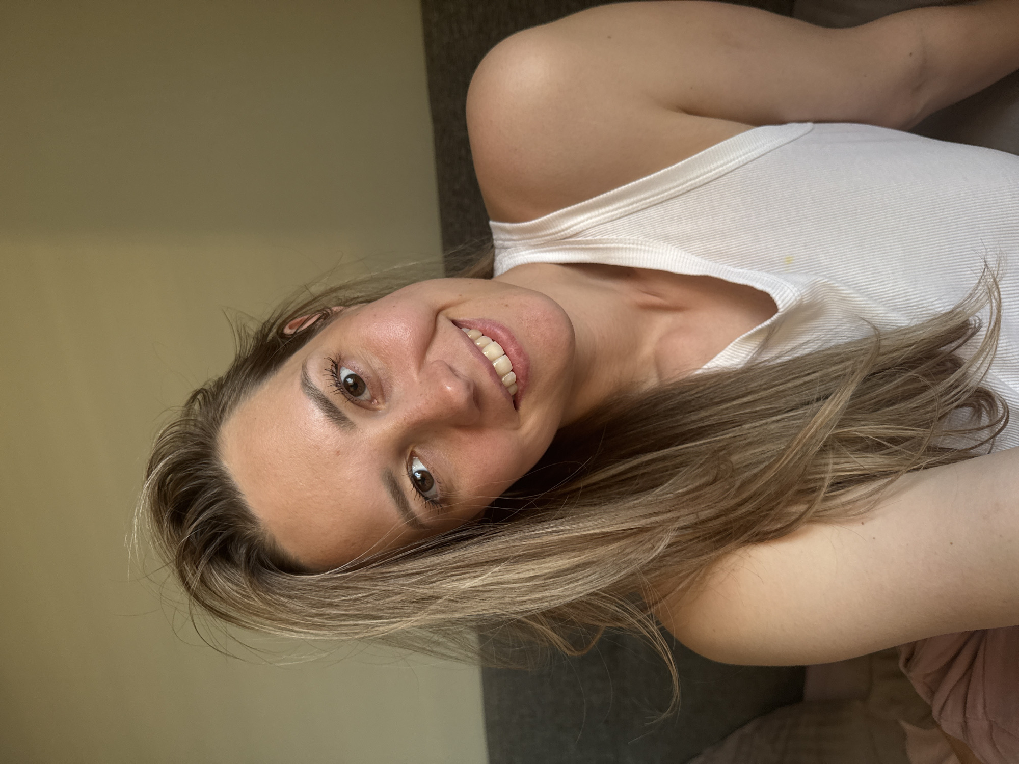A woman with light brown hair and brown eyes smiles at the camera while lying down. She is wearing a white tank top. The background is a beige wall and a gray couch. There is no evidence to suggest this photo was taken in Chaikovsky, Russia.