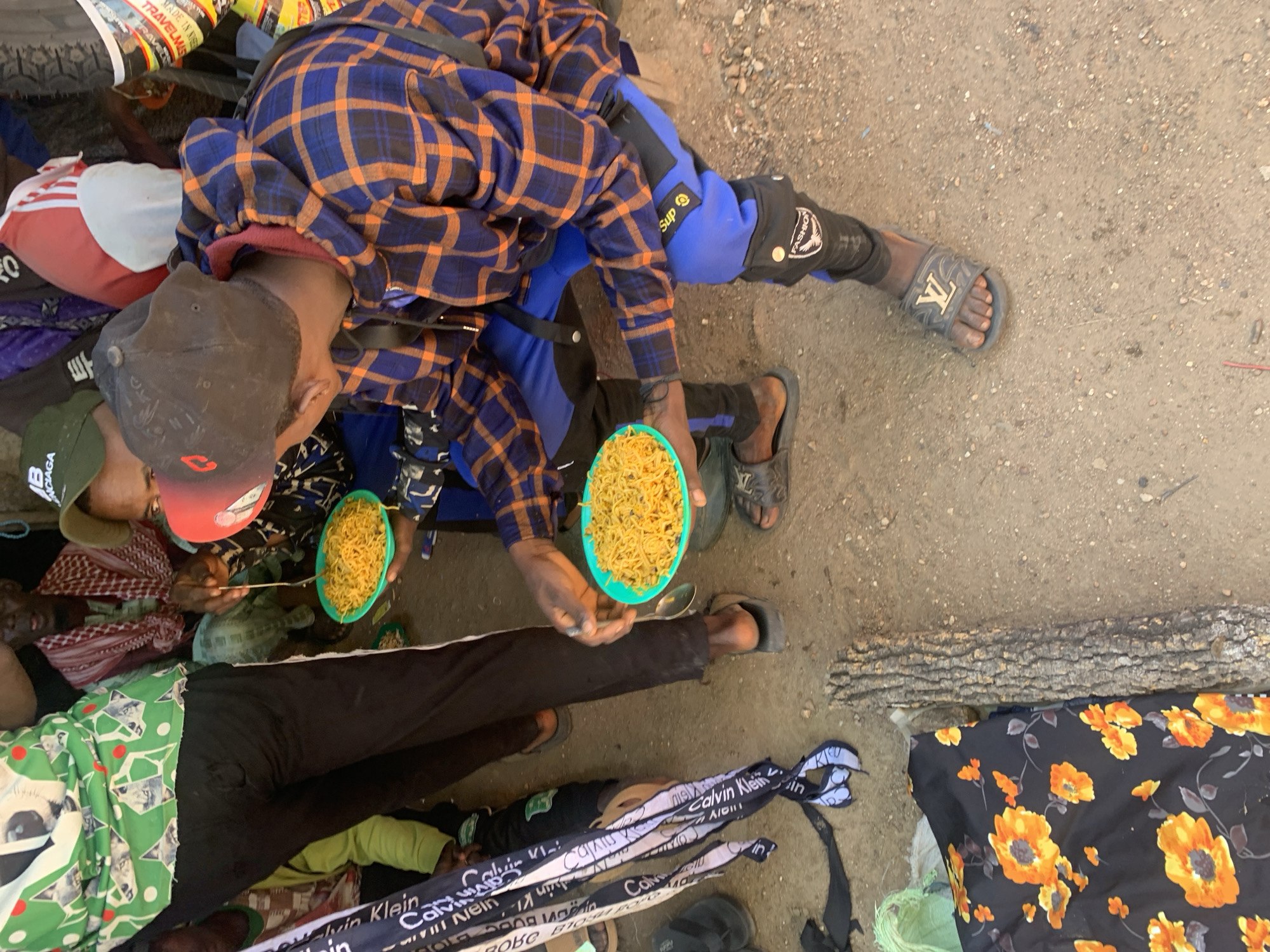 This media file depicts a group of people in what appears to be an outdoor, dusty setting, likely in Boi A, Nigeria. The subjects are clustered together on the ground, with some lying down and others seated or kneeling. One individual, wearing a blue and orange plaid shirt and blue pants with "Sup" and "Fashion" text, is holding a teal-colored bowl of noodles and a spoon, seemingly in the process of eating or offering the food. Another person, wearing a dark cap and a patterned garment, is also in close proximity to the food.
The scene suggests a moment of communal gathering, possibly a mealtime or a break. The ground is covered in dirt and small stones, indicating an unpaved area. Several individuals are covered by blankets or fabrics with various patterns, including a green and white geometric design and a black fabric with orange floral prints. Additionally, there are pieces of cloth with "Calvin Klein" text visible. The overall atmosphere appears informal and perhaps even a bit crowded. The lighting suggests it could be daytime, with natural light illuminating the scene. There are no strong indicators of specific weather conditions, but the dusty environment might imply dry weather.