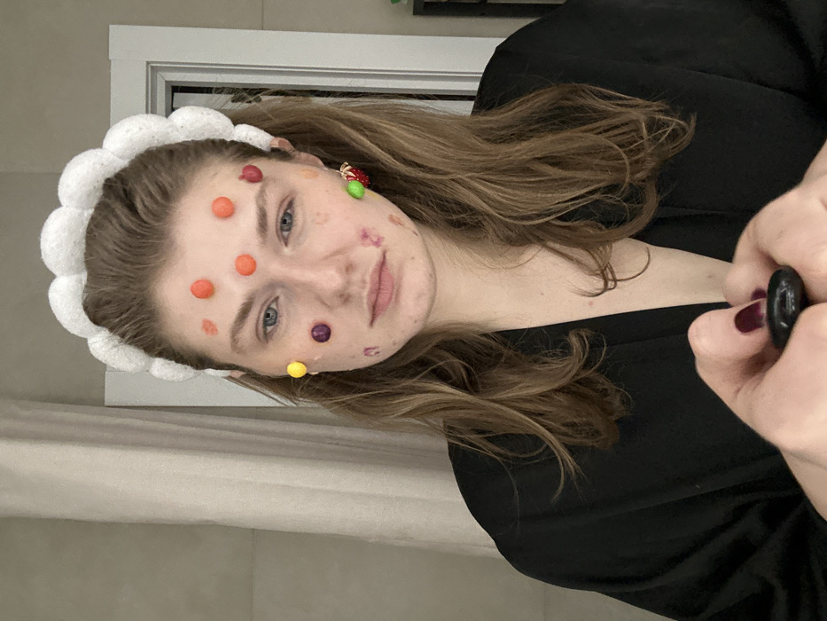 An individual is depicted indoors in what appears to be a residential setting within the city of Kazan, Russia. The subject is a female with light brown hair, appearing to be in her early twenties, facing directly towards the viewer with a neutral facial expression. She wears a white, scalloped, fluffy headband and a dark-colored robe or garment. Numerous small, brightly colored spherical candies, including orange, red, yellow, and purple variations, are adhered to her forehead, temples, and cheeks. A distinct strawberry-shaped earring, red with green accents, is visible on her left ear. Her fingernails are painted a dark, possibly burgundy, color. Both hands are positioned at the bottom center of the frame, holding a small, smooth, dark, oval-shaped object. The background features a light-colored curtain on the left and a white door frame on the right.