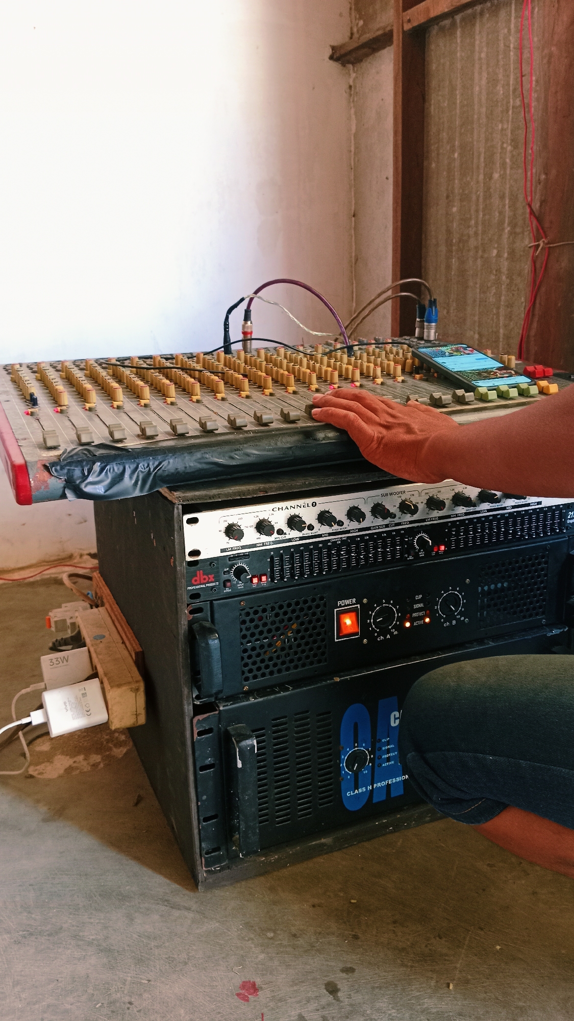 In a dimly lit, rustic room, a hand rests on a large, multi-channel audio mixer. The mixer, showing signs of use, is covered in faders and knobs. Cables, including purple and white ones, are connected to the mixer. Beneath the mixer, a rack-mounted system of audio equipment is visible. The top unit has controls labeled "CHANNEL 1" and "SUB WOOFER," along with smaller knobs and faders. Below it, a black unit labeled "dbx PROFESSIONAL PRODUCTS" has a red power indicator and several smaller lights. The bottom unit is a power amplifier, black with a large grille and a glowing orange power button. A smartphone is placed on the mixer, displaying content. On the left side of the image, near the floor, a white mobile phone charger labeled "33W" is plugged into a power strip. The environment appears to be a storage or working area, with rough concrete flooring and walls that appear to be plaster and corrugated metal. The overall impression is one of a functional, albeit somewhat cluttered, audio setup. The presence of audio equipment suggests a location likely used for events, performances, or recording within the city of Karang Indah, Indonesia, possibly in a community hall, a small venue, or a workshop.