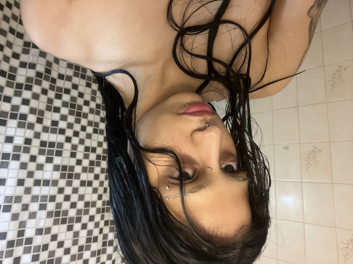 A person with dark, wet hair looks directly at the camera. Multiple facial piercings are visible above the eyebrow, on the nose, and on the lips. The person is fair-skinned. A black and white checkered wall appears on the left, contrasting with a wall of light-colored tiles on the right. The image orientation is rotated, making the individual appear upside down.