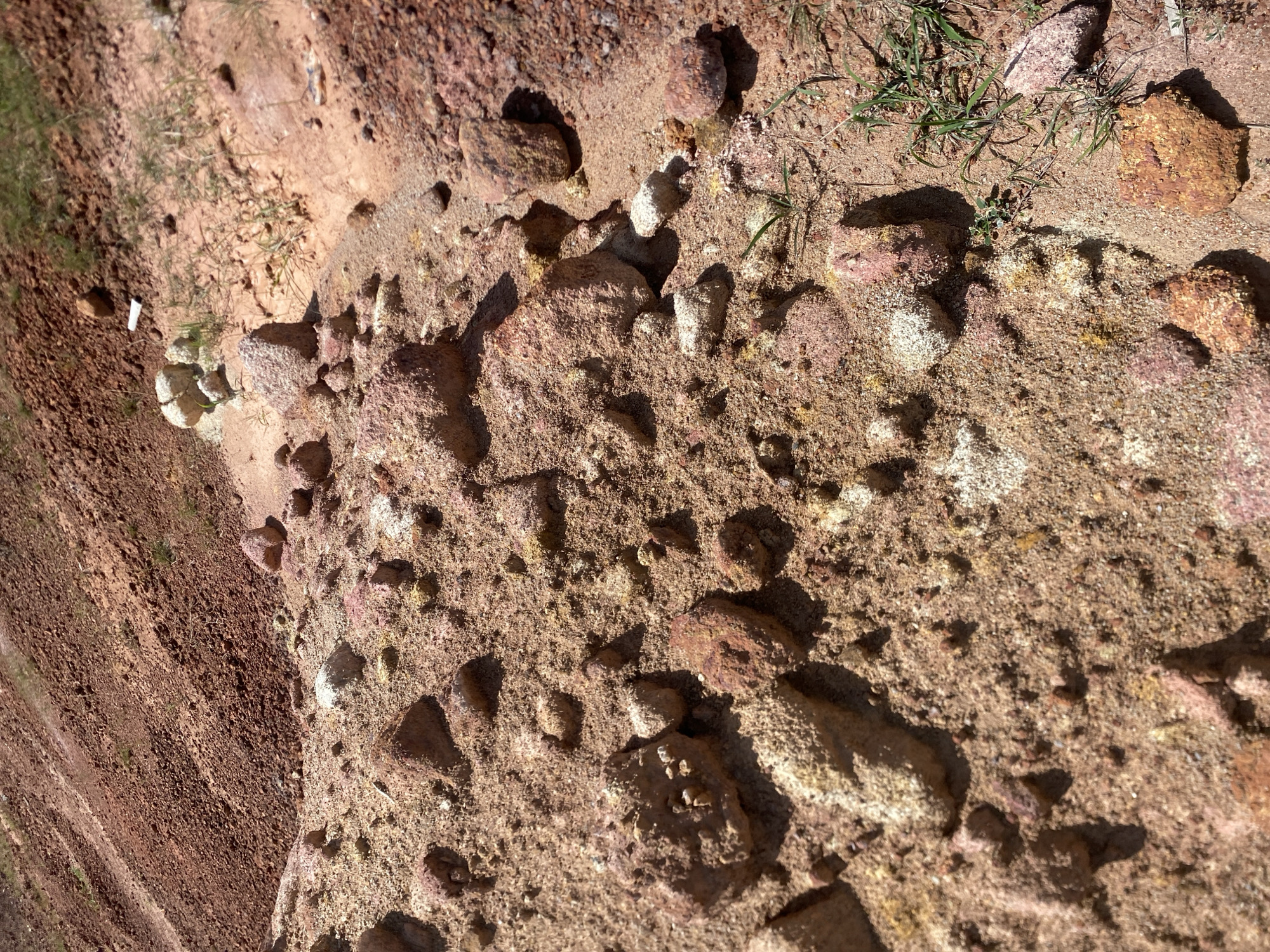 The image displays a close-up, ground-level view of a reddish-brown, sandy terrain with scattered rocks and sparse green vegetation. The landscape appears to be a hillside or embankment, with variations in soil composition and texture evident. Larger, rough-textured rocks are interspersed with finer sand and smaller pebbles. Patches of light-colored, possibly mineralized, deposits are visible on some of the rocks and in the soil. The lighting suggests a bright, sunny day, casting shadows that highlight the contours of the terrain. No people or discernible man-made structures are present. The scene is consistent with a natural, undeveloped area, possibly a construction site or erosion-prone zone.