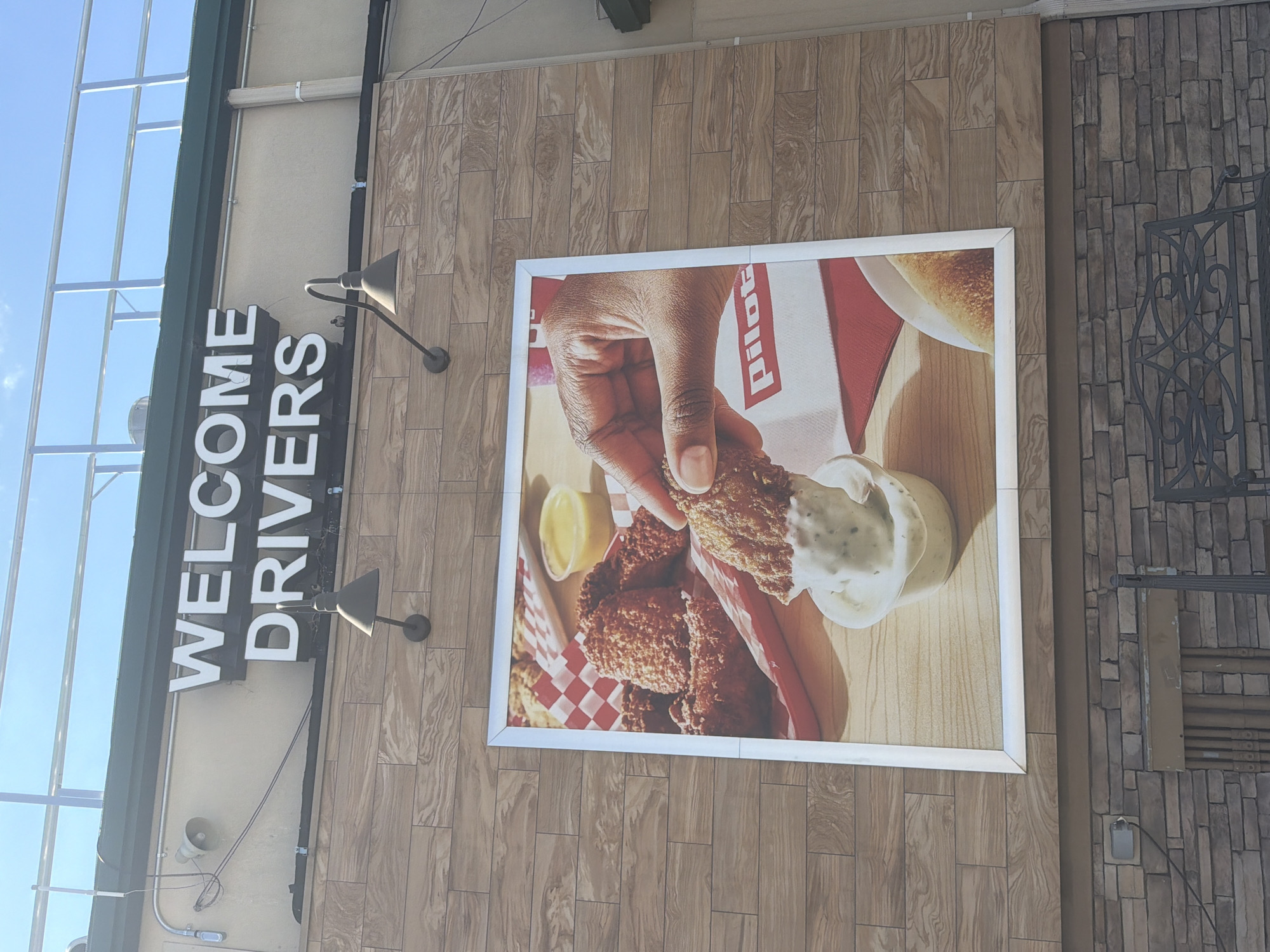 The image shows a sign that reads "WELCOME DRIVERS" above a large advertisement featuring fried chicken. The advertisement depicts a hand dipping a piece of fried chicken into a dipping sauce, with more chicken and a small cup of sauce in the background. The building has a facade of wood-like tiles, and below that, a stone facade. A metal bench is visible in the foreground. The overall scene appears to be an exterior view of a food establishment. The location is identified as the city of Lebanon, United States of America.