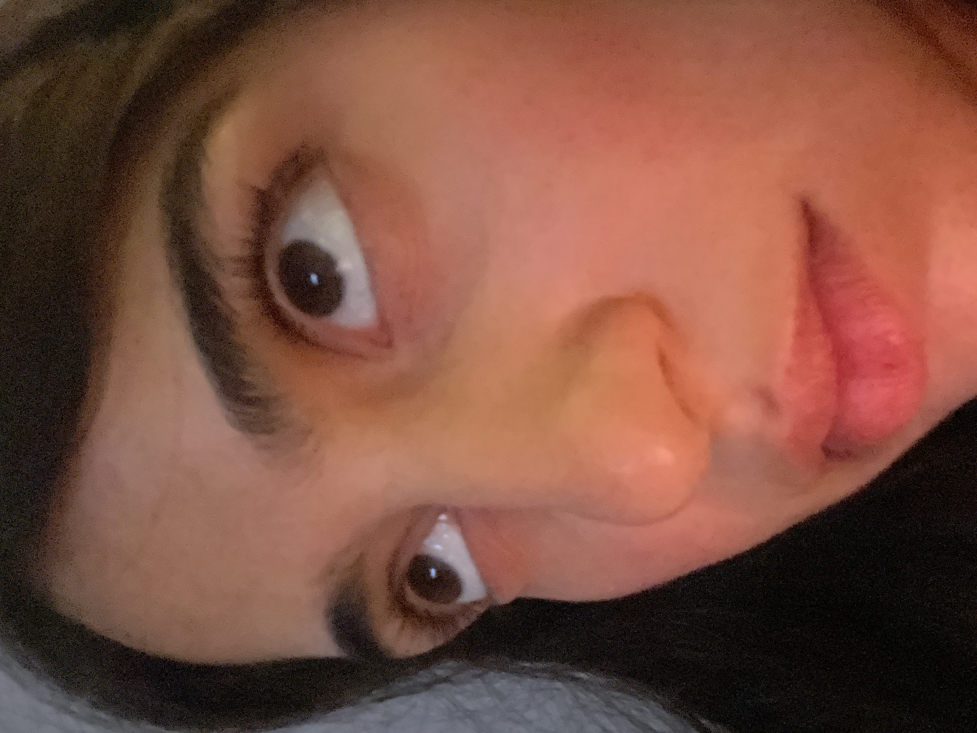 A close-up portrait of a person's face, slightly rotated clockwise. The visible features include the eyes, eyebrows, nose, and lips. The person has dark hair. The background is out of focus and indistinct. The location and city are unknown.