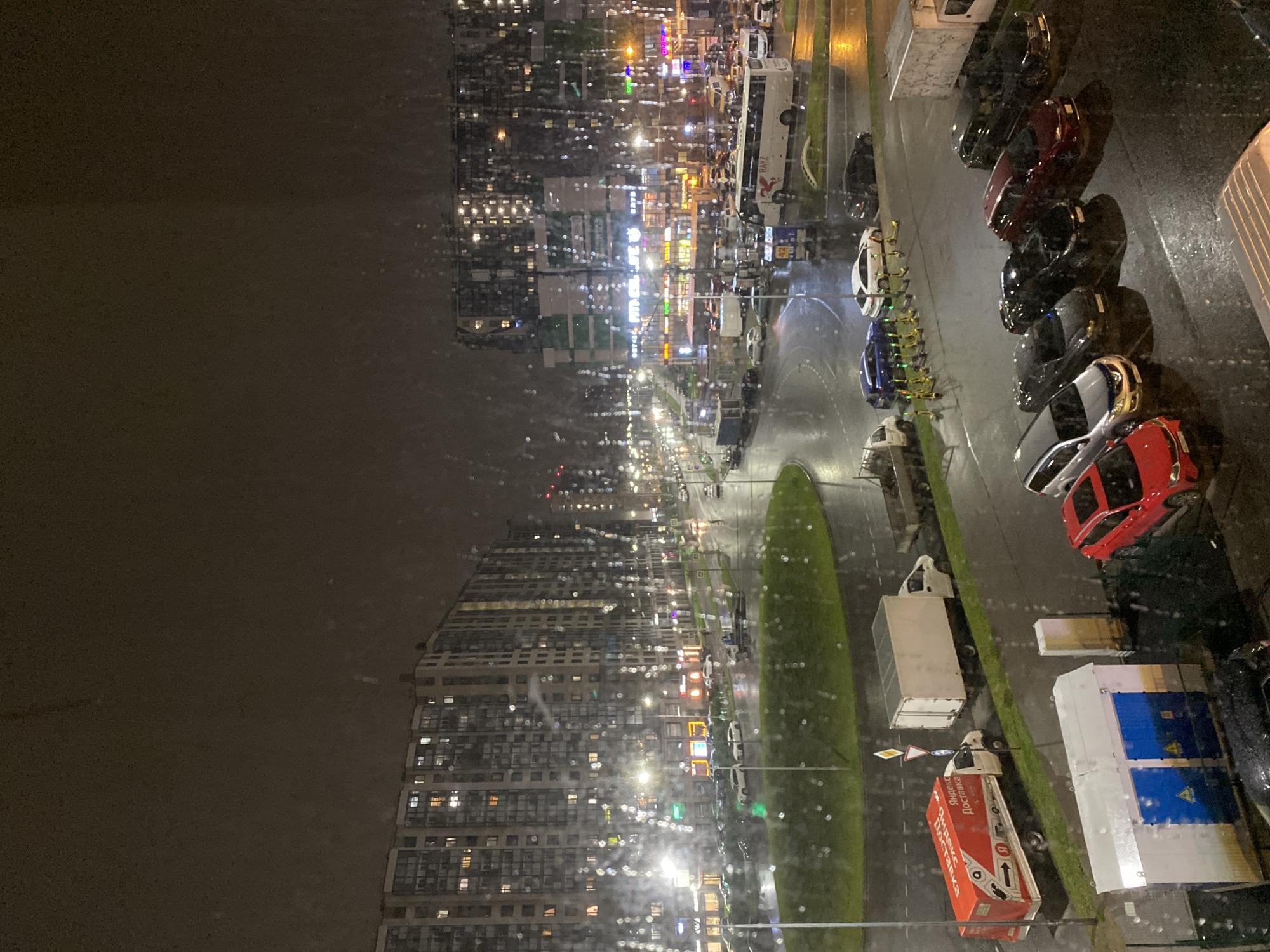 Here's a description of the image:
The image depicts a night view of an urban scene, likely from a high vantage point, possibly a window. Raindrops are visible on the glass, partially obscuring the view.
On the right side of the image, a row of parked cars is visible, including red, black and gray models. Adjacent to the cars, there are also several delivery trucks parked, including one with an "Яндекс Доставка" logo.
The central part of the image shows a wide street with streetlights and buildings densely packed with many illuminated windows. This extends to the far background. A grassy median is visible along the street.
The overall environment appears to be a rainy evening in a busy urban area.