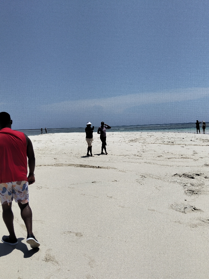 The image depicts a beach scene in Wasini, Kenya, under a clear blue sky. In the foreground, a person is observed from the rear, walking away from the camera. This individual wears a red sleeveless shirt, white and orange patterned swim shorts, and dark athletic shoes. Multiple footprints are visible in the light-colored sand.
In the mid-ground, two individuals are standing on the beach. The person on the left wears a white hat, a dark long-sleeved top, and light-colored shorts; they are holding a white object, likely a smartphone, elevated as if capturing an image or video. The person to their right wears a dark top and dark shorts, with one hand raised near their head.
Further in the background, towards the right, two additional figures are visible near the water's edge. One appears to be holding a dark object up, possibly a camera or phone, while the other stands beside them. In the far background, to the left, two indistinct figures are visible closer to the waterline. The ocean, characterized by light blue water, stretches across the horizon. The scene is brightly lit by natural sunlight.
