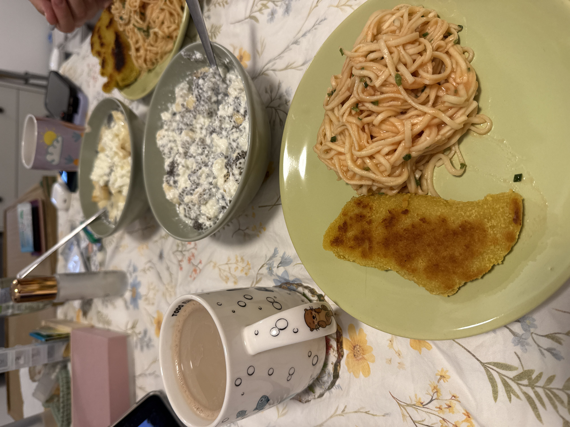 The image shows a table set with a meal. There's a light floral tablecloth with plates and bowls of food. On a green plate, there's pasta with a reddish sauce and a fried piece of what appears to be polenta. Other bowls contain what looks like cottage cheese and another type of mixed dish. A mug filled with a light brown liquid is in the foreground. The location is potentially in City of Zagreb, Croatia, based on the user's request.