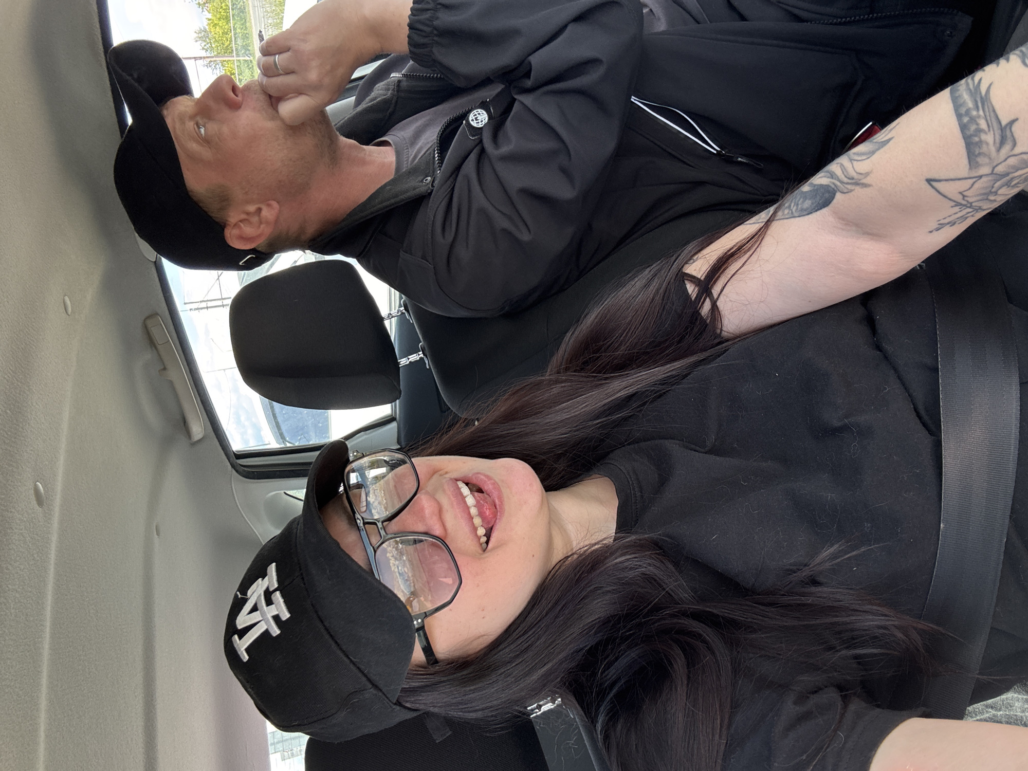 Two people are inside a car. A woman wearing a black shirt, glasses, and a black cap with "LA" on it is in the foreground. She is smiling with her teeth visible. Behind her, a man in a black jacket and black cap is holding his fingers to his mouth. A tattoo is visible on the man's arm. The car seats are black. Both are wearing seatbelts. The location is likely a car in the city of Unknown, Unknown.