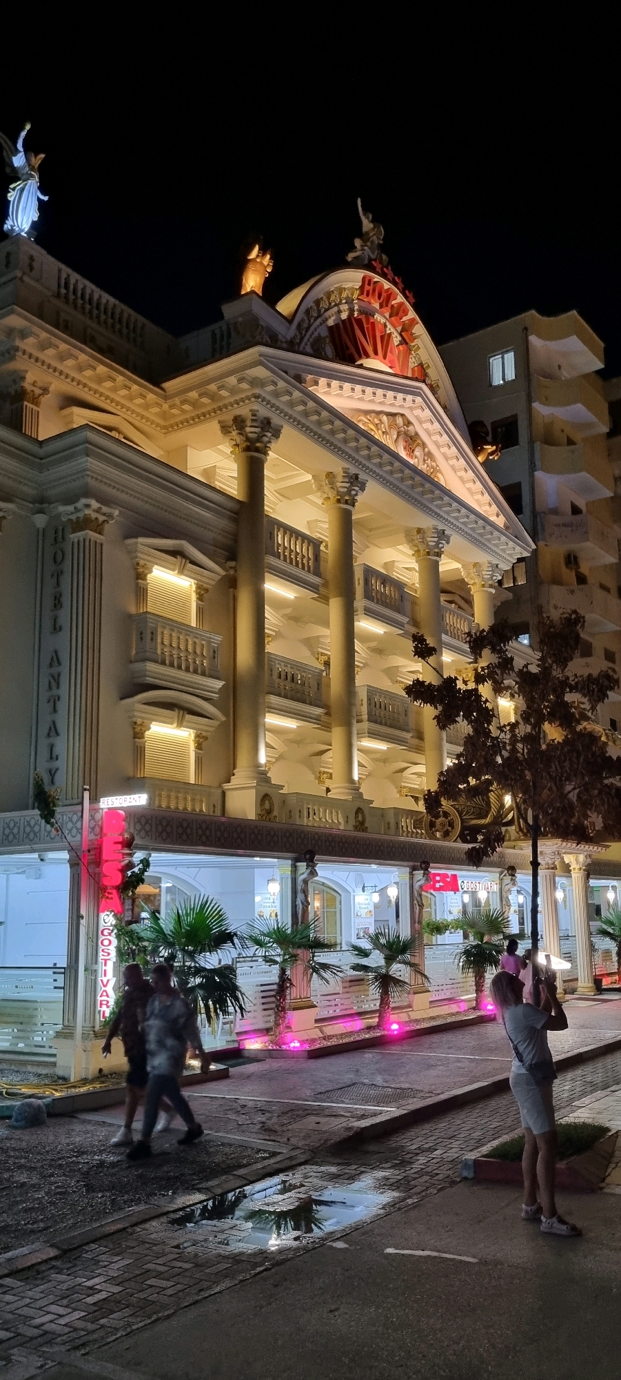 The image captures a nighttime scene in the city of Durrës, Albania, focusing on a lavishly lit hotel facade. The architecture is ornate, featuring classical columns, balconies, and decorative statues adorning the roof. The building is illuminated with warm, golden lights, highlighting its intricate details and creating a dramatic contrast against the dark sky. The hotel name "HOTEL ANTALY" is visible vertically along the left side of the building in white lettering. Below the main hotel structure, a restaurant area is visible with palm trees and vibrant pink lighting. A neon sign with red lettering reads "BESA GOSTIVARI". Two individuals are walking on the sidewalk in front of the restaurant, their figures blurred due to motion. Further to the right, a woman stands on the pavement, holding a phone up to take a picture of the hotel. The ground is wet, reflecting the lights and suggesting recent rain. In the background, a taller, modern apartment building stands in stark contrast to the classical style of the hotel.