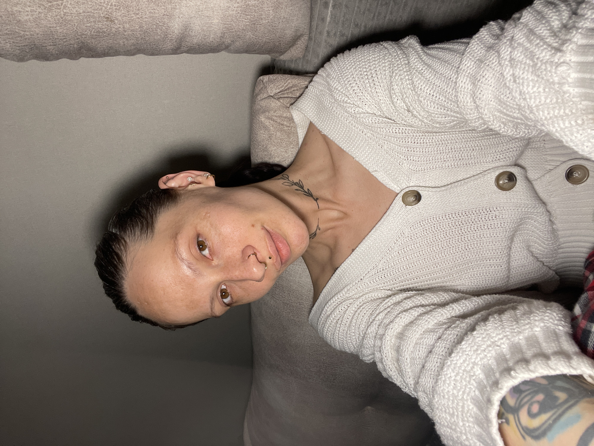 A close-up selfie shows a person with brown eyes and dark hair, pulled back, looking towards the camera. The image is rotated counter-clockwise. The person is indoors, likely resting on a light-colored couch or sofa. They are wearing a white, textured knit cardigan with two light brown buttons visible on the front.
Notable details include multiple piercings: several small stud and hoop earrings on the left ear, a heart-shaped septum piercing, and a small stud piercing above the upper lip (philtrum piercing). A delicate black tattoo of a leafy branch design is visible on the left side of the neck/collarbone area. Part of a larger, colorful tattoo is visible on the lower right arm, peeking out from under the cardigan sleeve. The person's expression is neutral or slightly contemplative. The lighting appears to be artificial, suggesting an indoor environment, possibly in the evening. There is no visible text in the image. The location context provided is Izhevsk, Russia.