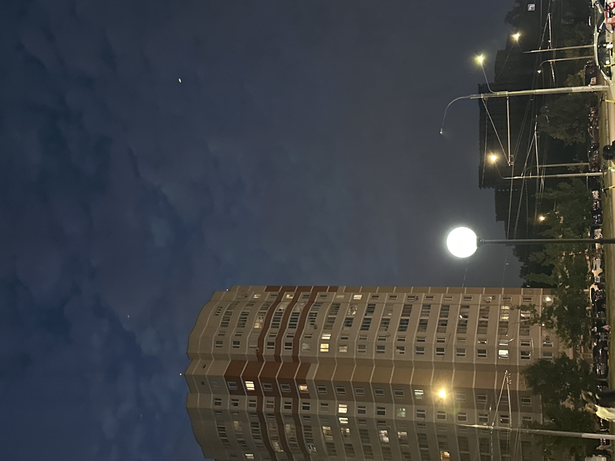 The image captures a nighttime street scene in Podolsk, Russia, dominated by a tall, light-colored apartment building with red and beige vertical stripes. The building has numerous windows, many of which are illuminated. In the foreground, a spherical street lamp with a bright light is visible. A second, less prominent street lamp illuminates the scene from the left. In the background, other buildings and trees are silhouetted against the dark sky, which features scattered clouds and two faint celestial bodies, possibly stars or the moon. Several cars are parked along the street, and a few people are walking or standing on the sidewalk. The overall impression is of a typical urban evening environment.