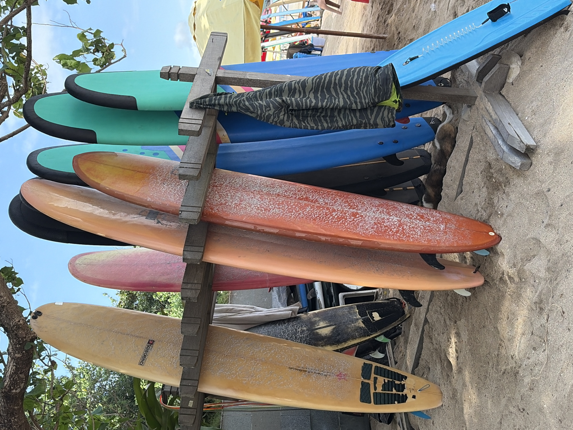 The image shows a rack of surfboards on a sandy beach in Kuta, Indonesia. The surfboards are of various colors and sizes, with some leaning against a wooden rack. A striped jacket is hanging from the rack. In the background, there are trees, a yellow umbrella, and other surfboards. A dog is lying on the sand near the surfboards. The sky is blue and clear.