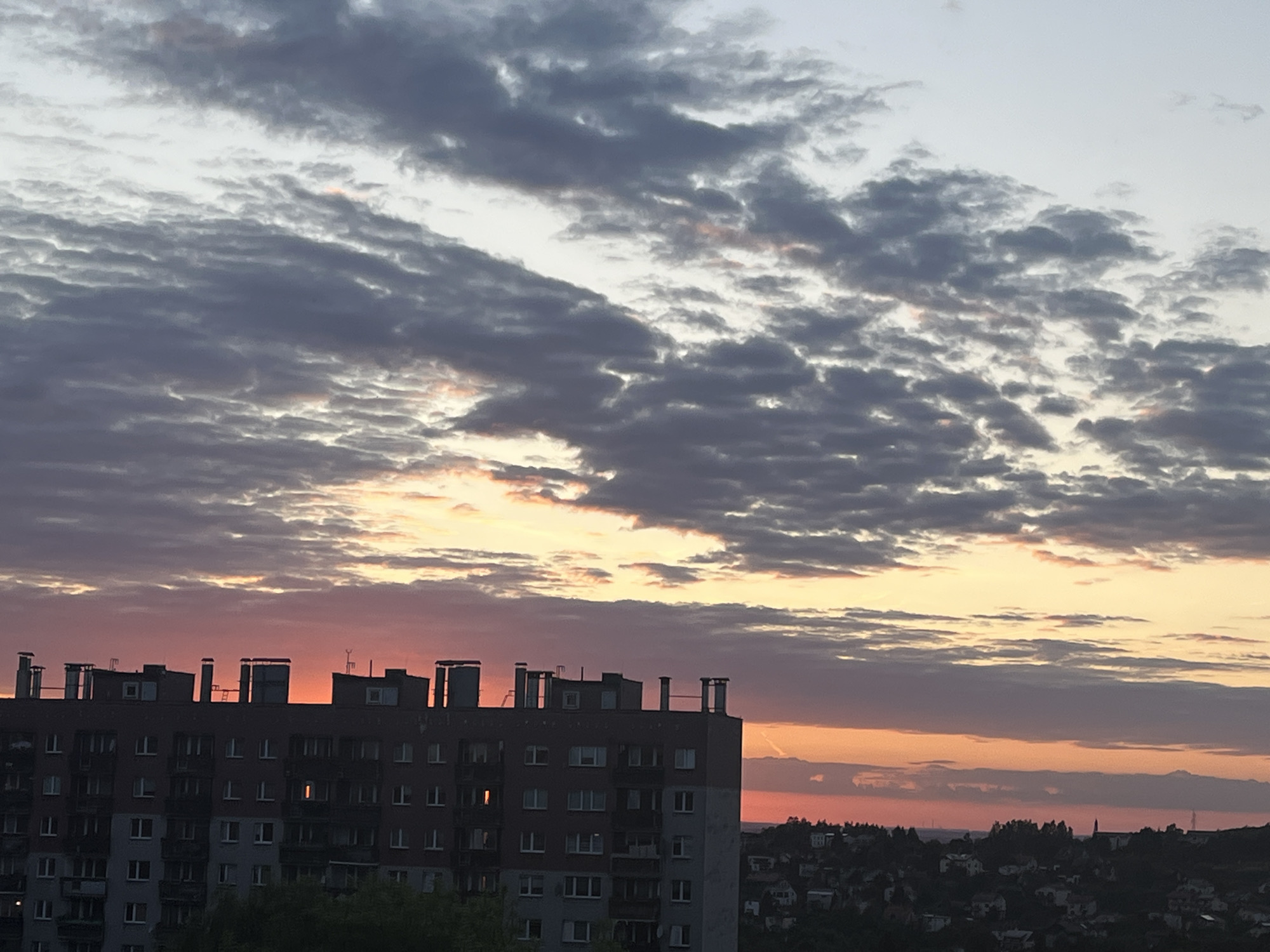 A wide shot captures a hazy sunset over a city. The sky is filled with clouds, tinged with pink, orange, and purple hues. In the foreground, a dark, multi-story apartment building dominates the left side of the frame. The building has a row of chimneys and ventilation systems on its roof. Below the building, the silhouette of a distant cityscape with scattered buildings and trees is visible. The overall impression is one of urban life at dusk.