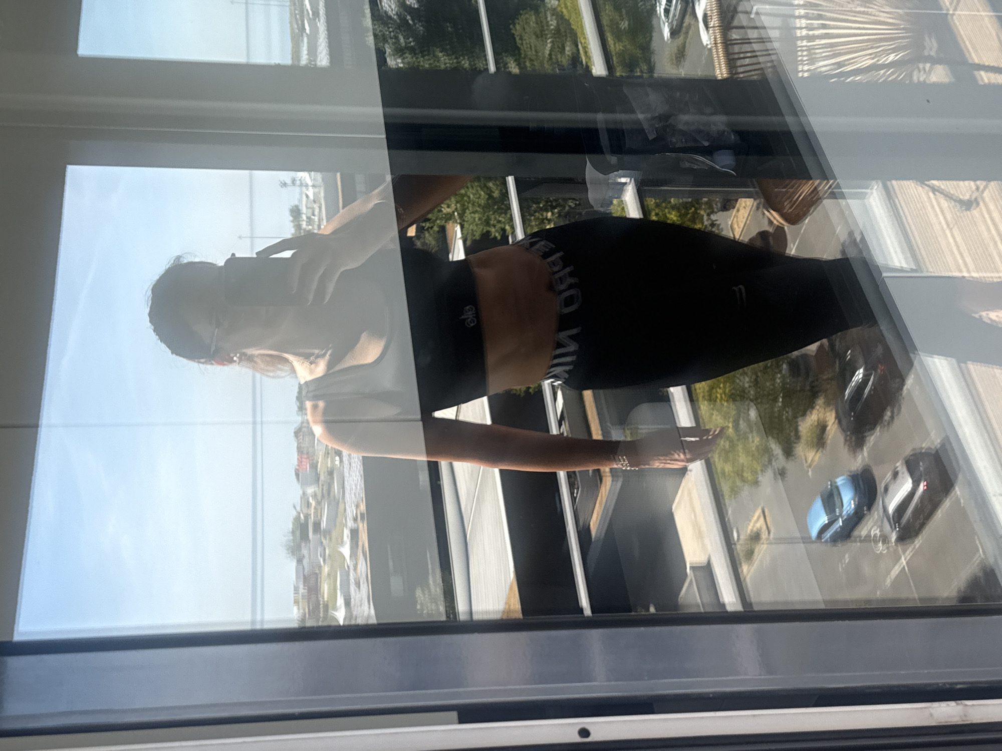 A woman is taking a mirror selfie with her phone on a balcony. She is wearing a black sports bra and black leggings. Her abdomen is visible and appears toned. The background shows a city view with buildings, trees, and cars. The image is taken from a reflection in a glass window, likely from a building in Loures, Portugal.