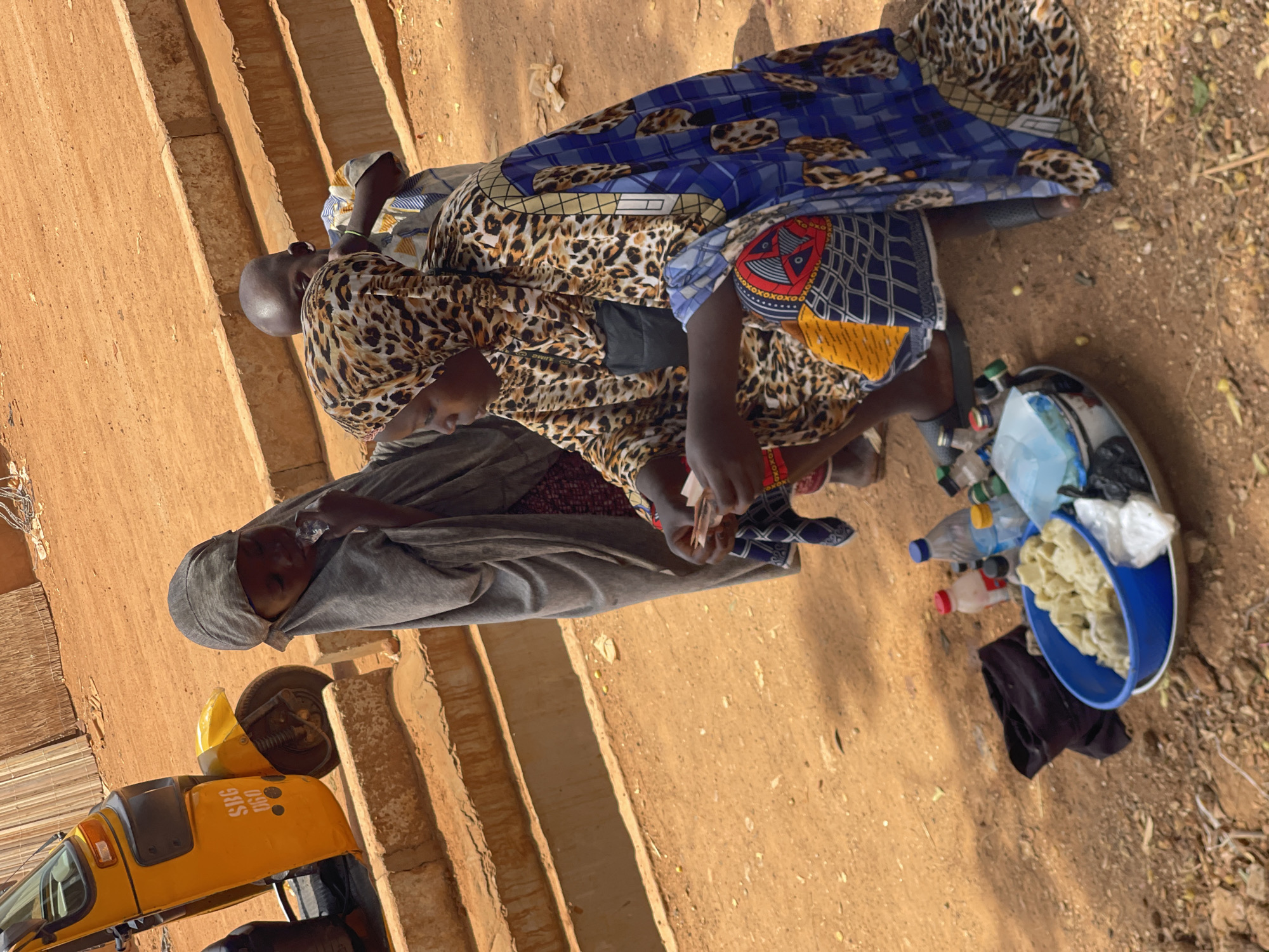 The image, correctly oriented, captures an outdoor scene in Zaria, Nigeria, featuring three women and a vehicle. The ground is unpaved and dusty, with earthy-toned steps or a raised platform visible in the background.
In the center, a woman wearing a leopard print top and a vibrant patterned skirt (featuring blue, yellow, red, and black designs) is engaged in an interaction. Her hands are visible, suggesting an exchange of items or money. Facing her from the left is another woman dressed in grey attire with a matching grey head covering. A third woman, also in a leopard print and blue outfit, is partially visible behind the central figure.
On the ground to the right, a blue plastic basin contains various items, including white, possibly food-related products (such as pastries or dough), and several plastic bottles. A piece of black fabric rests beside the basin.
In the left background, a yellow motorized rickshaw, characteristic of local transportation, is parked. The text "SJG 060" is visible on its side. The scene appears to be a common daytime interaction, possibly a street vending or market transaction, under clear, sunny weather conditions.