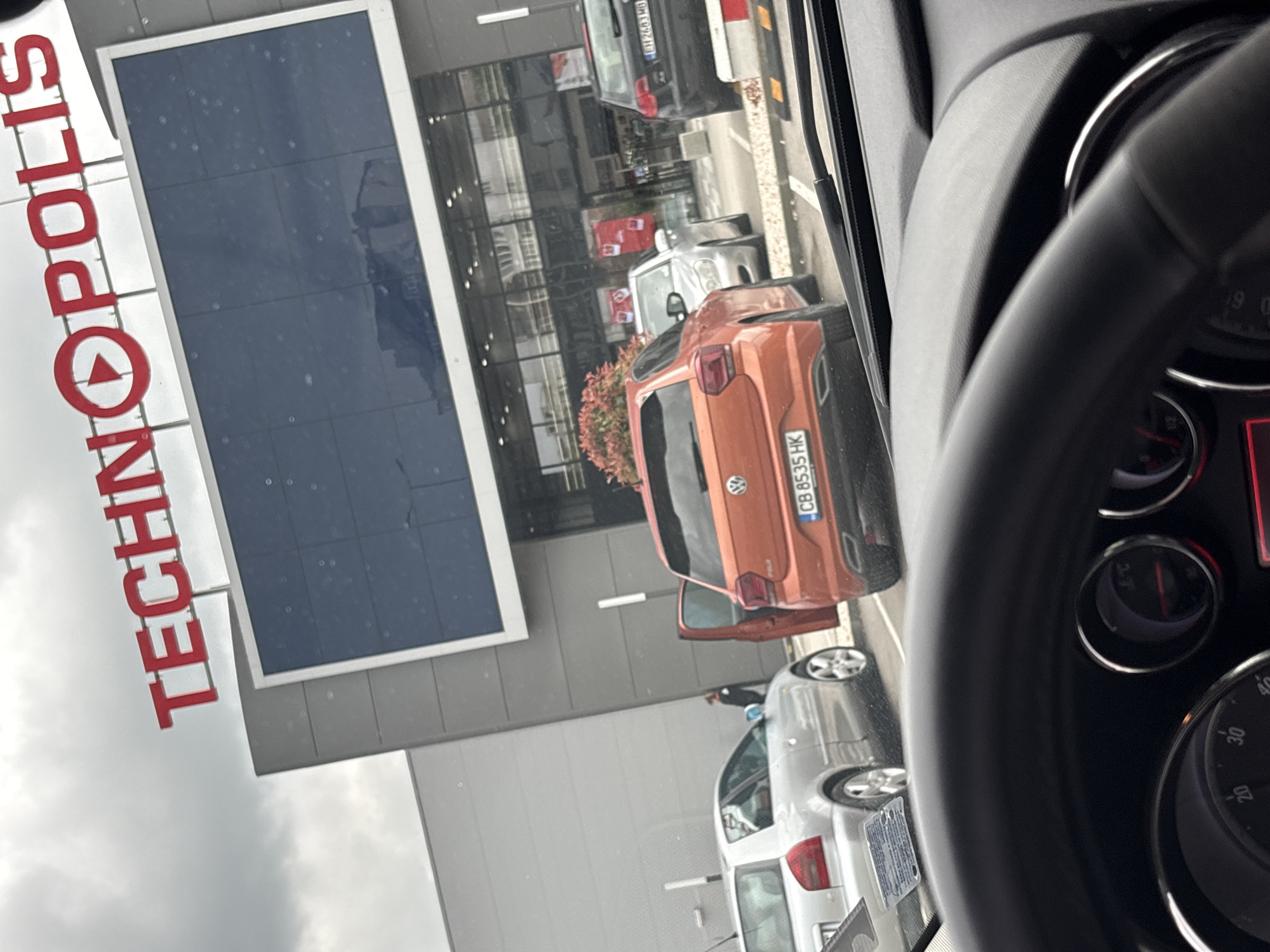 The image shows a view from inside a car, likely a driver's perspective, looking towards a TECHNOPOLIS store. The store is identifiable by its large sign, which is partially visible. In the foreground, there's a steering wheel.
The scene outside the car includes several parked cars, most notably a small orange Volkswagen with a license plate reading "CB 8535 HK". The building's architecture seems modern with large glass windows. The overall atmosphere seems urban, likely a commercial district.
The location is identified to be in Lovech, Bulgaria.