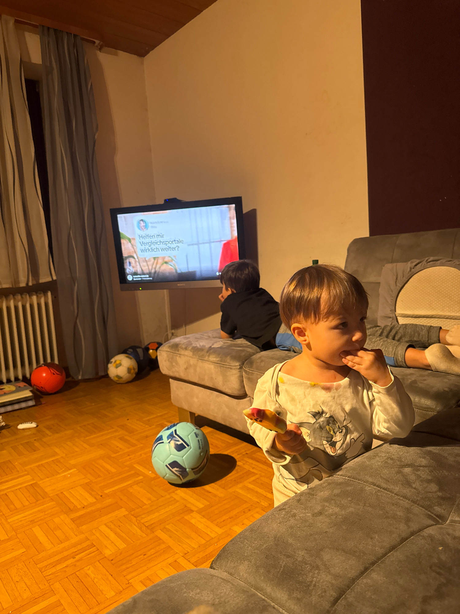 The image depicts an indoor scene, possibly a living room, with a television displaying text in German. Two young children are present. One child, in the foreground, is wearing a white long-sleeved shirt with a cartoon image of Tom the cat and is holding a colorful object to their mouth, possibly food. This child has light brown hair and is looking to the right. In the background, another child, with dark hair, is seated on a grey sofa facing the television. Several soccer balls are scattered on the wooden floor near the sofa, along with a stack of books. A radiator is visible behind a curtain on the left side of the image. The lighting suggests it is evening or indoors with artificial light. The image does not provide any specific information about the location within Überherrn, Germany.