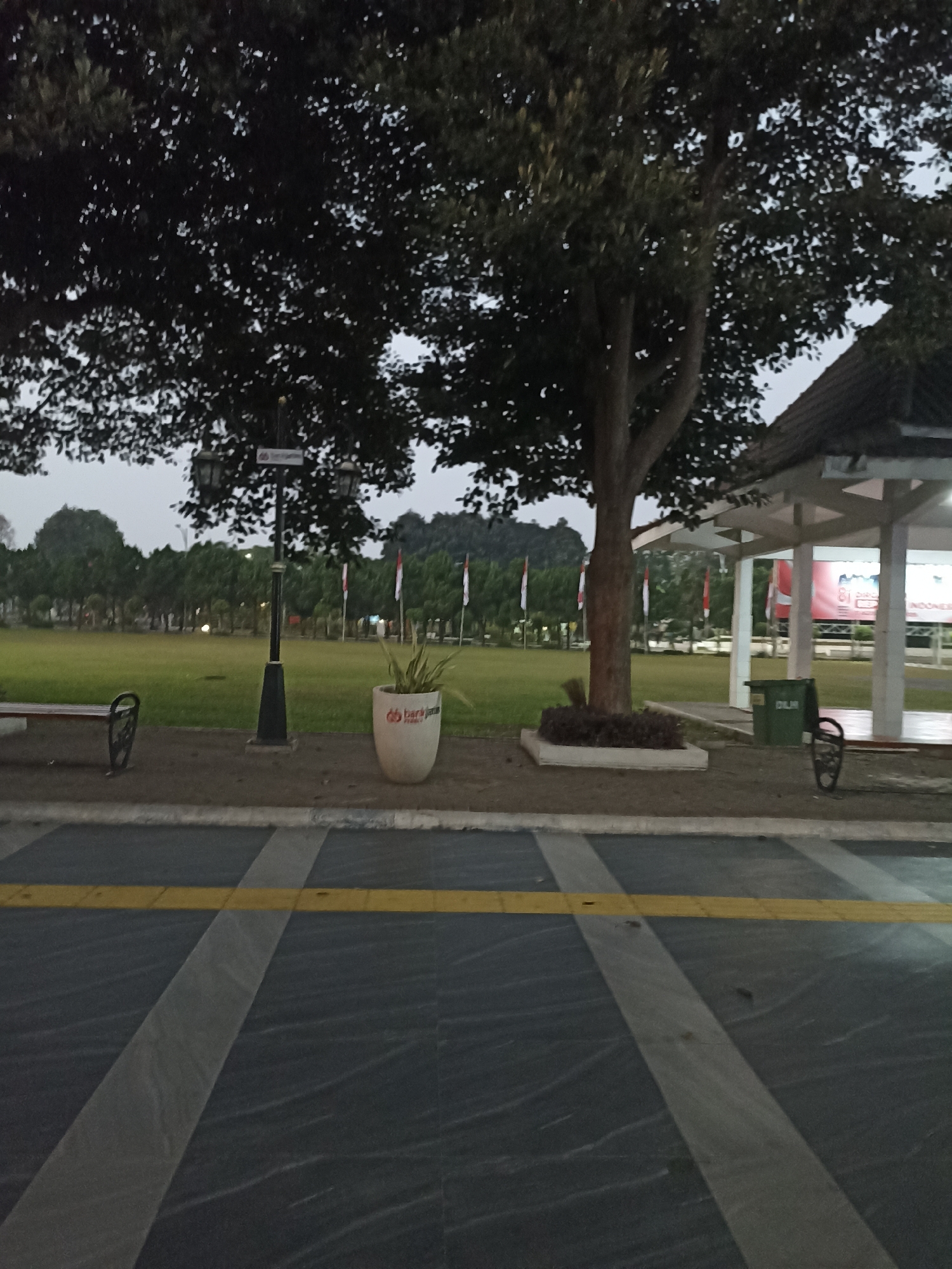 The image depicts an outdoor scene, likely a public park or plaza, in Lumajang, Indonesia. In the foreground, a dark asphalt surface with light grey and yellow striped markings is visible. Moving further back, a paved walkway separates the asphalt from a grassy field. Along the walkway, there are two dark benches. To the right of the walkway, a large white planter with a potted plant is situated. A black lamppost with two lanterns stands near the center of the walkway. Beyond the walkway is a vast green field, bordered by a line of trees in the middle ground. Several Indonesian flags on poles are visible throughout the field. In the background, a white pavilion-like structure with a brown roof is present, and a green trash bin is next to it. The overall lighting suggests it is either early morning or late evening, with a cloudy sky. No people are visible in the image.