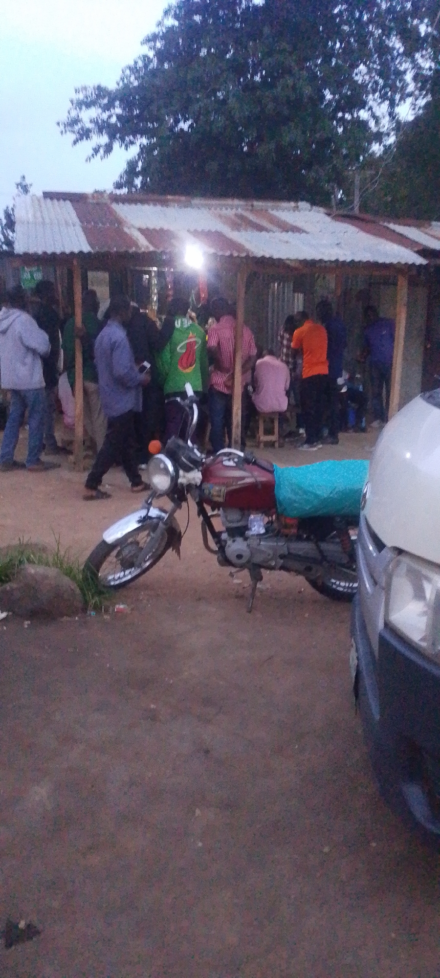 A group of people are gathered under a weathered, corrugated metal roof shelter, which appears to be a roadside structure. The shelter is supported by wooden poles and has a tin roof. Several individuals are standing and interacting within the sheltered area. A red motorcycle with a blue tarp strapped to its rear is parked in front of the shelter, partially obscuring the ground. To the right of the motorcycle, the front portion of a light-colored vehicle is visible, suggesting it is parked adjacent to the scene. The ground is a dirt or gravel surface. The overall lighting suggests it might be dusk or an overcast day. This scene is located in Gura Top, Nigeria.