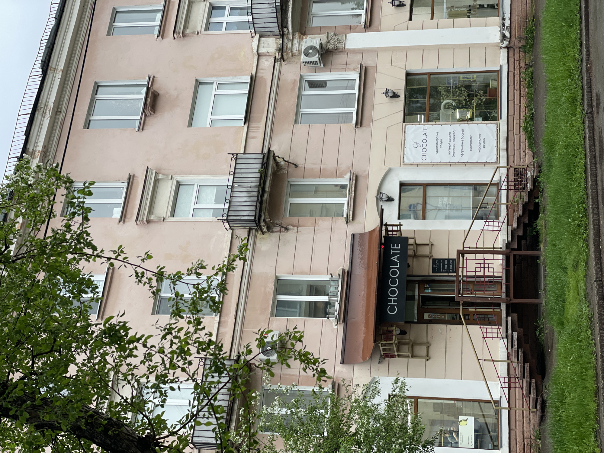 The image shows a multi-story building in Zheleznogorsk, Russia. The building is painted in a light pink color and features white-framed windows, some with small balconies. A sign above the entrance reads "CHOCOLATE" vertically, indicating a business located on the ground floor. The sign below the windows reads “CHOCOLATE” and lists salon services. There are metal stairs leading to the entrance. The building is partially obscured by tree branches with green leaves. The setting appears to be a street with grass and pavement visible on the right side.