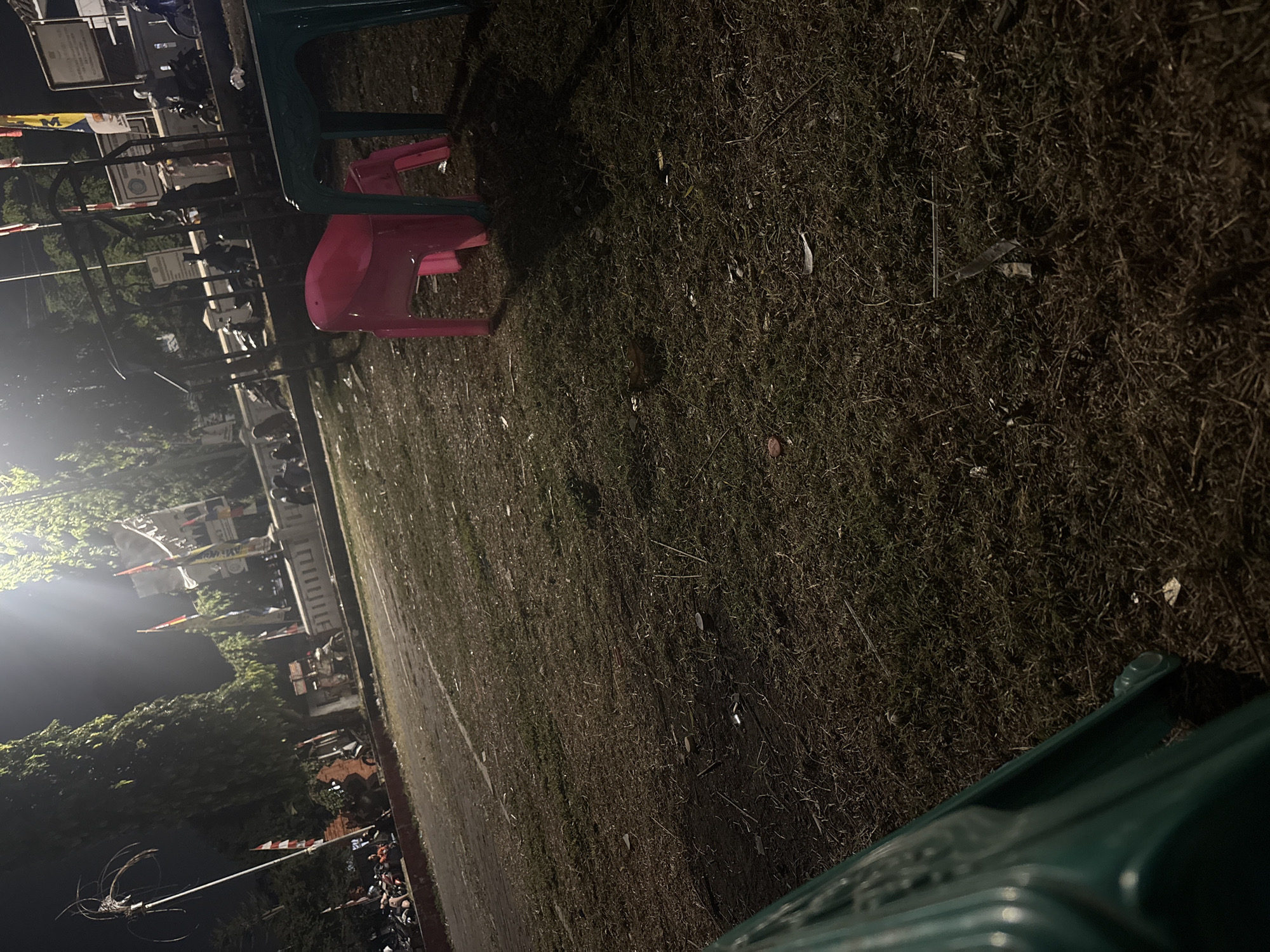 This image depicts an outdoor scene at night, likely in Palimanan, Indonesia. In the foreground, a section of a green plastic chair is visible. Further back, on the grass, a pink plastic chair is positioned. The ground is covered in dry grass and scattered debris. Beyond this area, a paved path is visible, with numerous motorcycles parked along its edge. Several people are present, some standing and others sitting, near a raised concrete structure adorned with flags and banners. Trees line the background, and bright lights illuminate the area, creating strong shadows. The overall impression is of a public gathering or event taking place after dark.