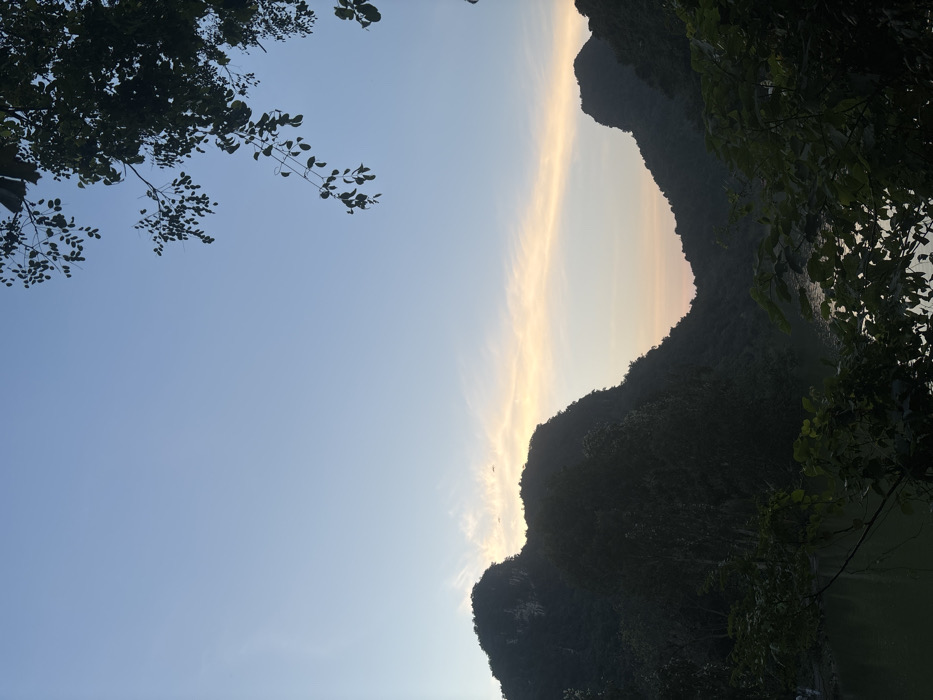 The image displays a natural landscape in Yên Sơn Ward, Vietnam, likely captured during either dawn or dusk. The sky transitions from a clear, light blue overhead to a pale orange and pinkish-white horizontal cloud band across the horizon. Dark, densely vegetated karst mountains form prominent silhouettes against the sky, framing a lower central area. A body of dark green water, consistent with a lake or river, is visible in the lower left and central portions, partially obscured by dark green foreground foliage. A tree branch with dark leaves extends into the upper right corner, while additional dense foliage occupies the lower right. Two small, dark, unidentifiable aerial objects are present in the sky above the left mountain. No human figures, structures, or artificial objects are discernible.