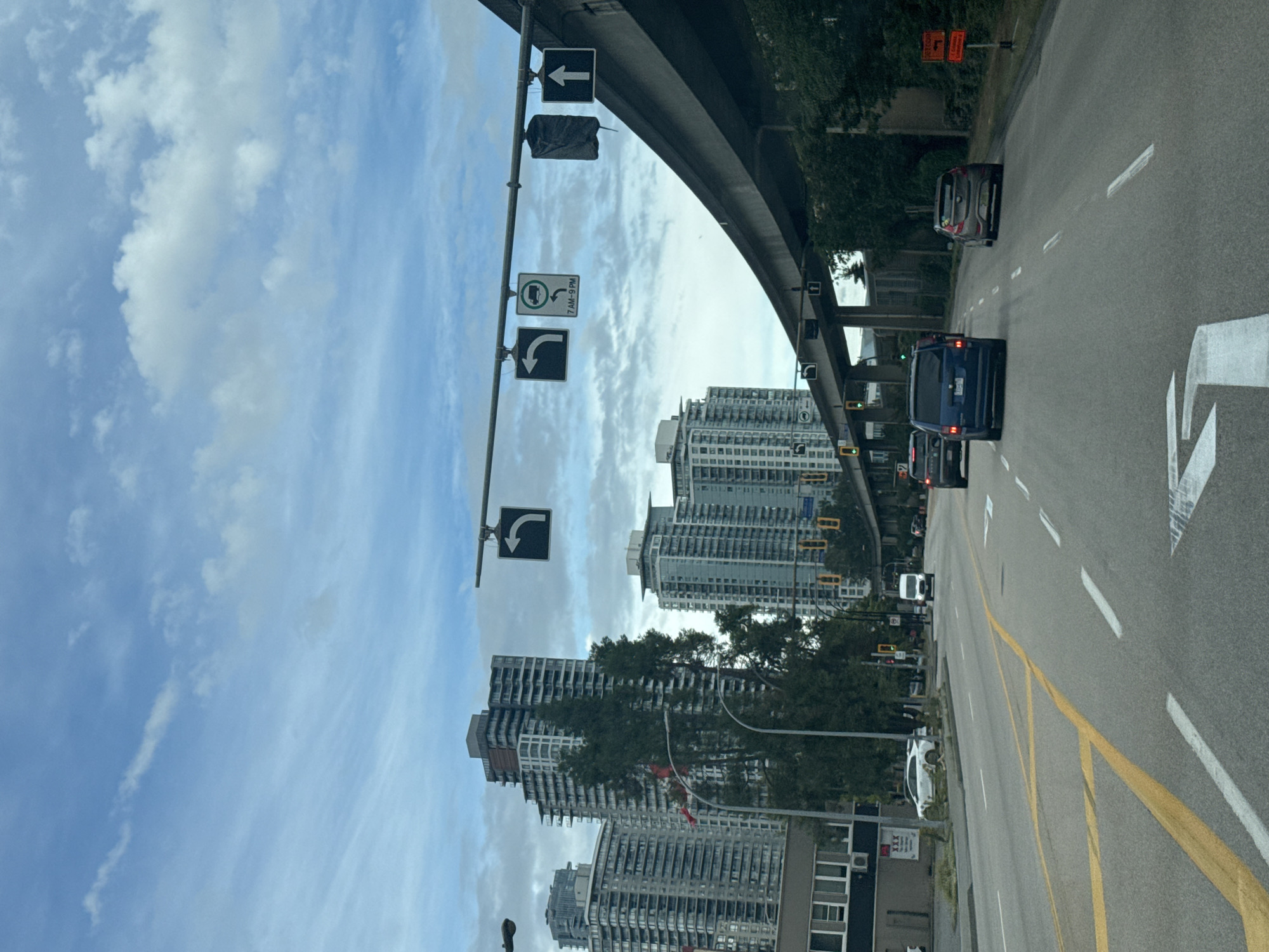 The image depicts a street-level view of a multi-lane road in New Westminster, Canada. Tall residential buildings are visible in the background, with modern architecture featuring many balconies. A raised, elevated roadway or transit line runs diagonally across the upper right portion of the image, supported by concrete pillars. Traffic signals are present, indicating a controlled intersection. Several vehicles are on the road, including cars and a van, traveling in the same direction as the viewer. Road markings, including yellow and white lines, delineate the lanes. The sky is partly cloudy. The overall environment suggests an urban setting with ongoing traffic flow.