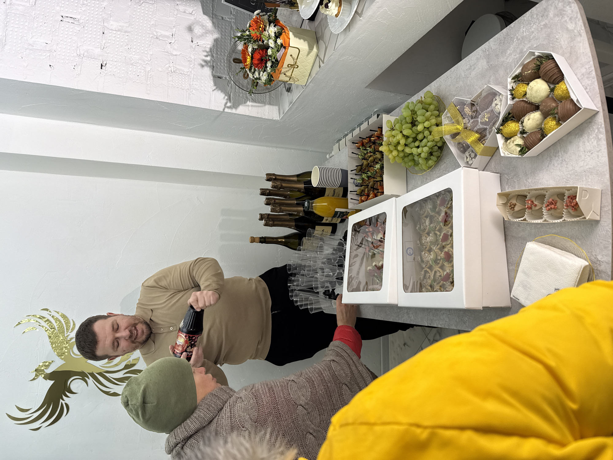 A man in a tan shirt is holding a bottle and talking to a person in a green beanie and brown sweater in a shop in Chaikovsky, Russia. The bottle has Cyrillic text on the label, indicating it's a product sold in Russia. On the counter next to them are stacked wine bottles, champagne flutes, boxes of desserts, grapes, and savory snacks. A bouquet of flowers and another dessert are on a shelf above the counter. A golden phoenix emblem is visible on the wall behind the man. Another person wearing a yellow jacket partially obstructs the view on the lower right side.