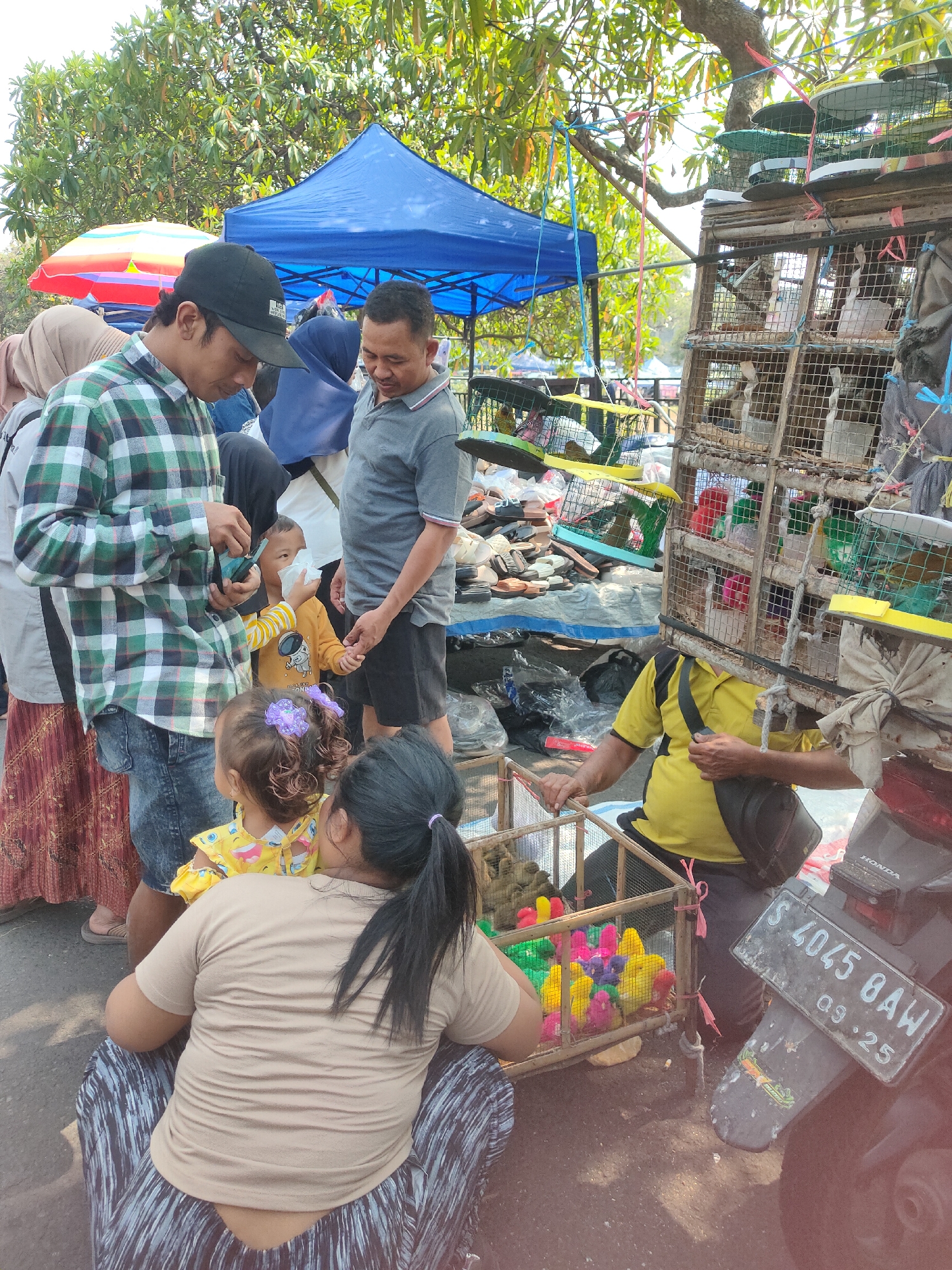In the city of Ngasem, Indonesia, an outdoor market scene unfolds beneath the dappled sunlight filtering through trees. In the foreground, a woman with black hair tied in a ponytail sits facing away from the camera, her back to the viewer, wearing a tan t-shirt and patterned pants. Next to her, a small child with curly hair and a purple clip in her hair, wearing a yellow shirt, looks towards the left. To their right, a toddler in a yellow shirt with a cartoon character is being held by a man in a plaid shirt and a black baseball cap. The man appears to be showing the toddler something on a small blue device he is holding. Beside them, another man in a gray polo shirt with black shorts stands and looks down.
Behind the people, a vendor with a yellow shirt and black accents, wearing a black bag across his chest, stands next to a large, multi-tiered wooden cage filled with birds. The cages are constructed from wood and wire mesh. In the background, more cages with birds are visible, suspended from wires and tree branches. A blue tent stands in the middle ground, and a colorful umbrella is partially visible to its left. On the ground, various items are laid out, possibly for sale, including what appears to be footwear and bags. A motorcycle with a license plate reading "S 4045 B4W 09.25" is parked to the right, partially obscuring some of the cages. The ground is dusty and uneven.