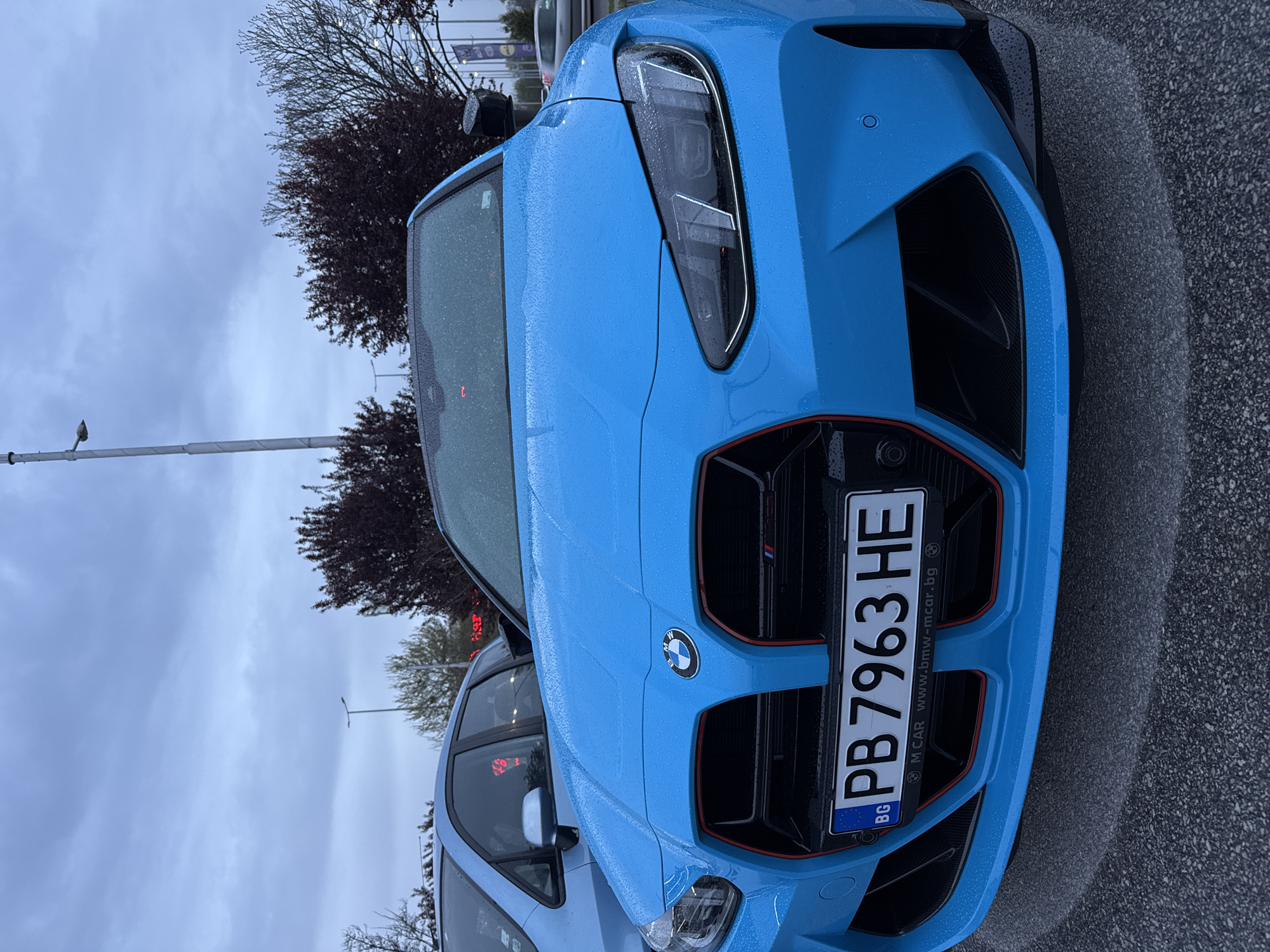 The image shows the front of a blue BMW car, partially covered in raindrops. The car has a Bulgarian license plate "PB 7963 HE." The background includes a tree, a lamppost, and cloudy sky. The car's license plate indicates the location is Bulgaria. Based on the license plate design and the website printed on it (www.bmw-mcar.bg), it is plausible the image was taken in Plovdiv, Bulgaria, as M Car, a BMW dealership is located there.