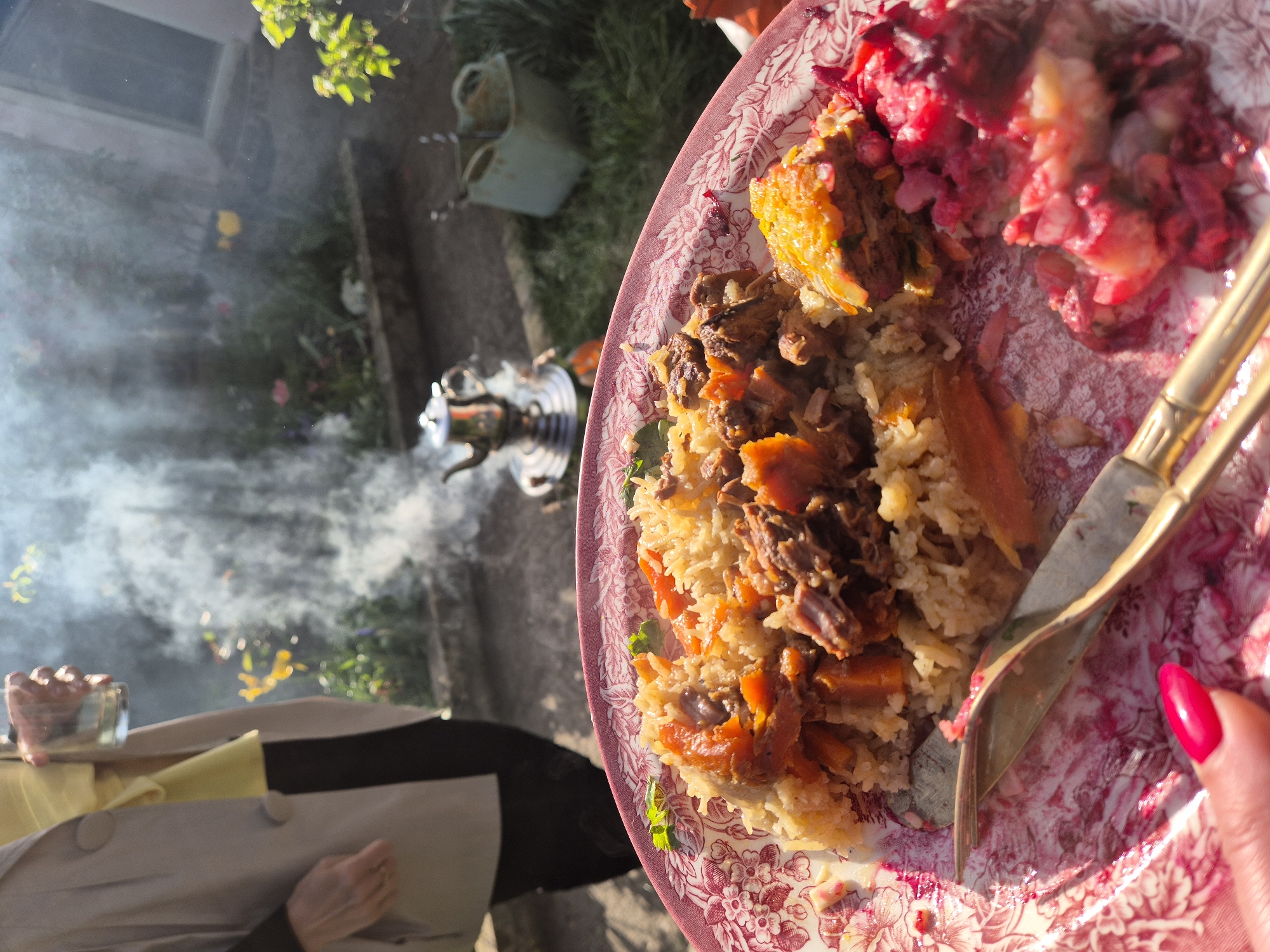The image shows an outdoor scene with a plate of food in the foreground. The plate contains a rice dish with meat and vegetables, and a beet salad. A fork and knife rest on the plate, and a hand with red nail polish is visible. In the background, there is a samovar, a person holding a glass, some greenery, and a building. There is smoke in the air. The location is unclear, and no landmarks suggest the city of Ipswich, United Kingdom.