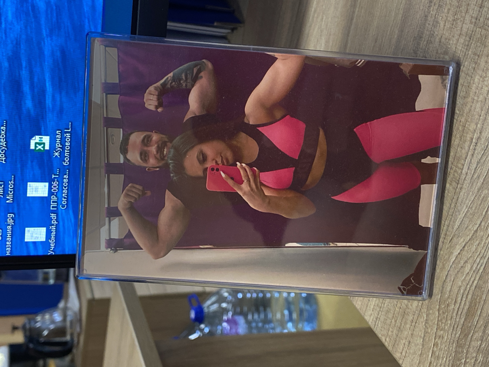 A photograph in a plastic frame rests on a wooden surface, possibly a table. The photo within the frame shows a couple flexing their muscles while posing for a selfie. The woman is holding a pink smartphone. The man has tattoos on his left arm. The woman is wearing a pink and black sports bra and matching leggings. The setting is indoors, with a computer monitor visible behind the photo, displaying Russian text. A water bottle sits on the table as well. The location and city, Luzhitsy, Russia, cannot be determined from the image alone.