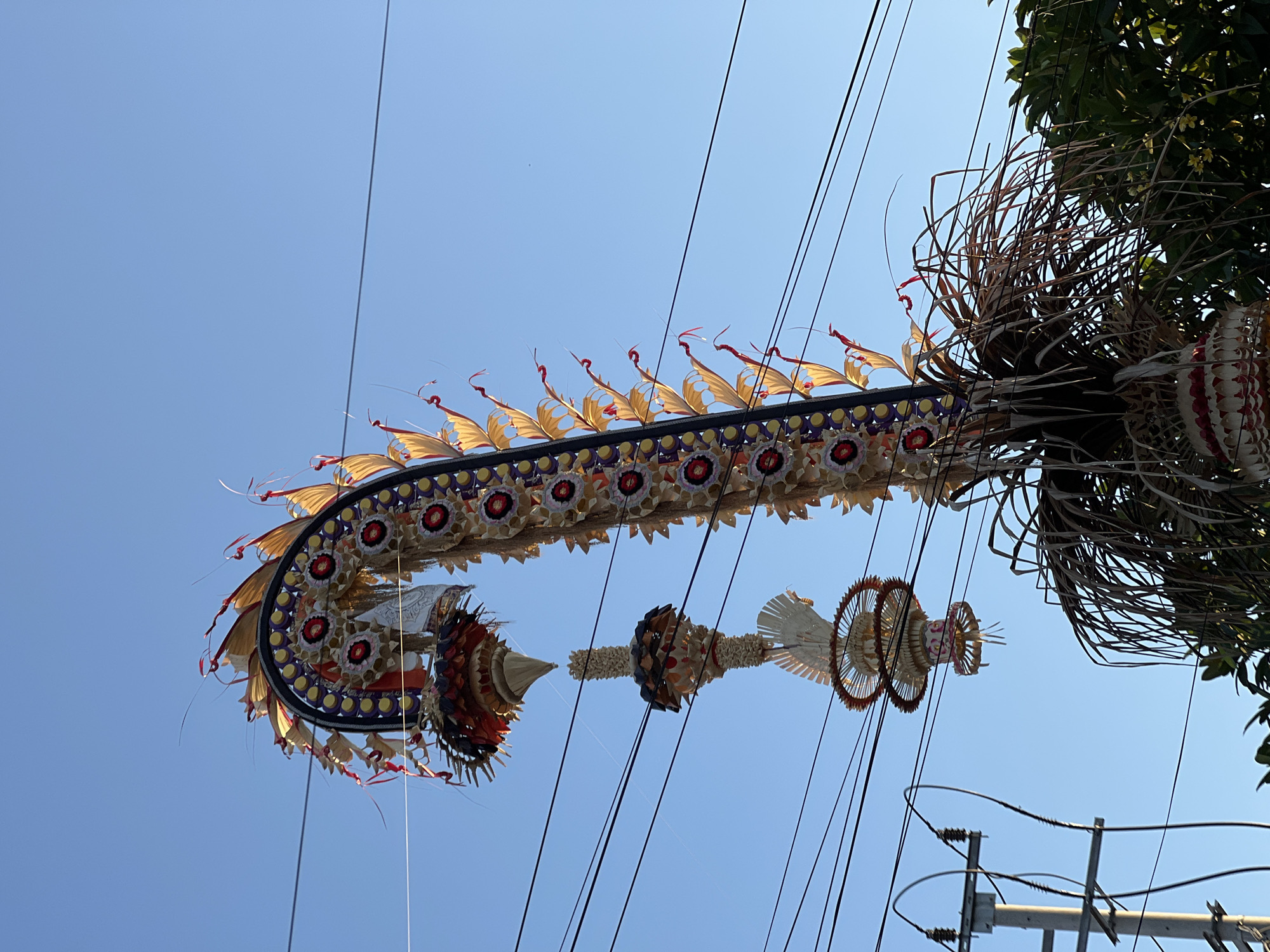 An ornate, festive structure, possibly a Penjor or similar Balinese decoration, stands against a clear blue sky in North Kuta, Indonesia. The structure is made of natural materials, including woven palm leaves, reeds, and colorful floral elements. It features a curved, arching top decorated with red and yellow petal-like shapes and circular motifs. Hanging below the main arch are several tiered, cone-shaped ornaments, each intricately woven and adorned. The entire structure is supported by a base of dense, dried grass-like material. Power lines crisscross the sky in front of the decoration, and a utility pole is visible on the left side of the frame. The image is taken from a low angle, looking upwards.