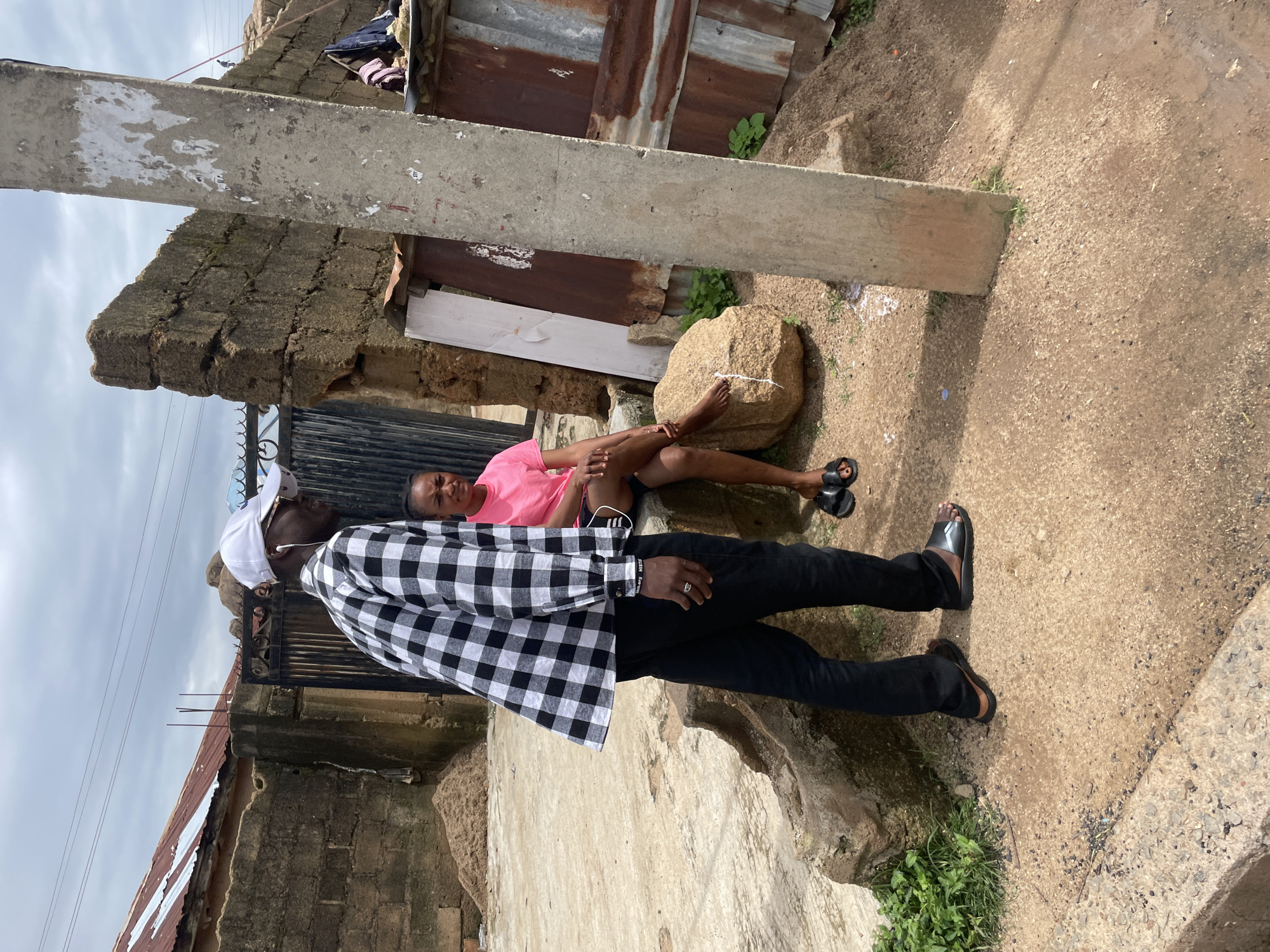 In Jos, Nigeria, a man wearing a black and white checkered shirt, black pants, a white cap, and sunglasses stands on a concrete path looking to his right. He has earbuds in and is wearing sandals. A woman in a pink t-shirt and black shorts is seated on a large rock to his right, also looking to her right. She is wearing black sandals. The background shows a rustic environment with stone walls, a metal gate, a corrugated metal structure, and a concrete pole. The ground is a mix of concrete and dirt, with some vegetation visible. The sky is overcast.